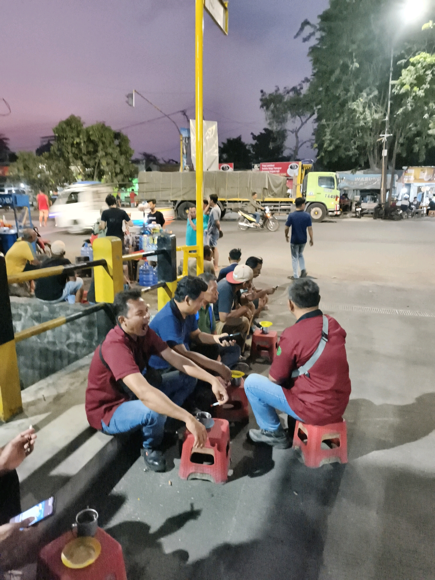 This image captures a group of men gathered outdoors in what appears to be a public space, possibly on the side of a road or in a communal gathering area. The setting is likely in Desa Ponokawan, Indonesia, given the context provided. The lighting suggests it is either dusk or early evening, with a purplish hue to the sky.
In the foreground, several men are seated on low, red plastic stools. They are casually dressed, with some wearing t-shirts and jeans. Some of the men are engaged in conversation, while others are smoking cigarettes. One man in a maroon t-shirt and blue jeans is prominently featured, looking towards the viewer with his mouth open. Another man in a blue t-shirt is also visible, seated beside him. In the immediate foreground, a hand holds a smartphone, and a cup sits on a small red stool.
Behind this group, there are more people, some standing and others sitting. A white van is parked nearby, and a yellow truck with the license plate "T 649" is also present, suggesting a roadside scene or a commercial area. Water bottles are stacked on a rack behind some of the seated men. Trees are visible in the background, and traffic infrastructure, including a yellow pole, is also part of the environment. The ground is paved with asphalt. The overall atmosphere appears to be relaxed and informal, with people gathered to socialize.