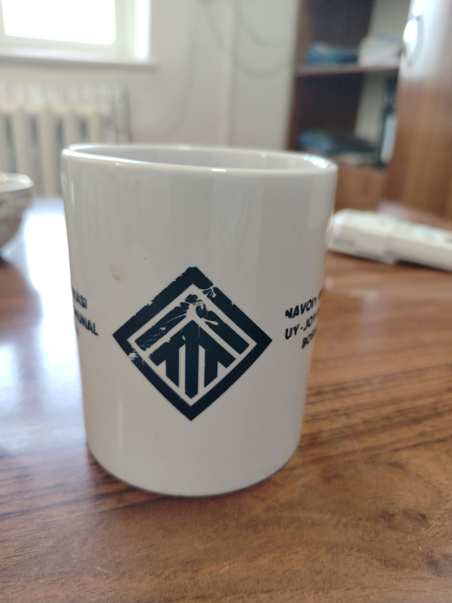 A white ceramic mug with a dark diamond-shaped logo containing an arrow design is placed on a wooden table. Text is visible on the mug, with "NASI" and "UNAL" on the left side, and "NAVOT" and "UY-JOY" on the right. A faint brown smudge is present on the mug to the left of the logo. In the background, a window with vertical blinds and a radiator are visible. A blurred wooden shelf unit is also present. A white electronic device, possibly a remote control, is on the table to the right of the mug. The scene appears to be indoors, in a domestic or office setting. There are no people visible in the image. The lighting suggests it is daytime. The location in Nurata, Uzbekistan cannot be determined from the visual information present.