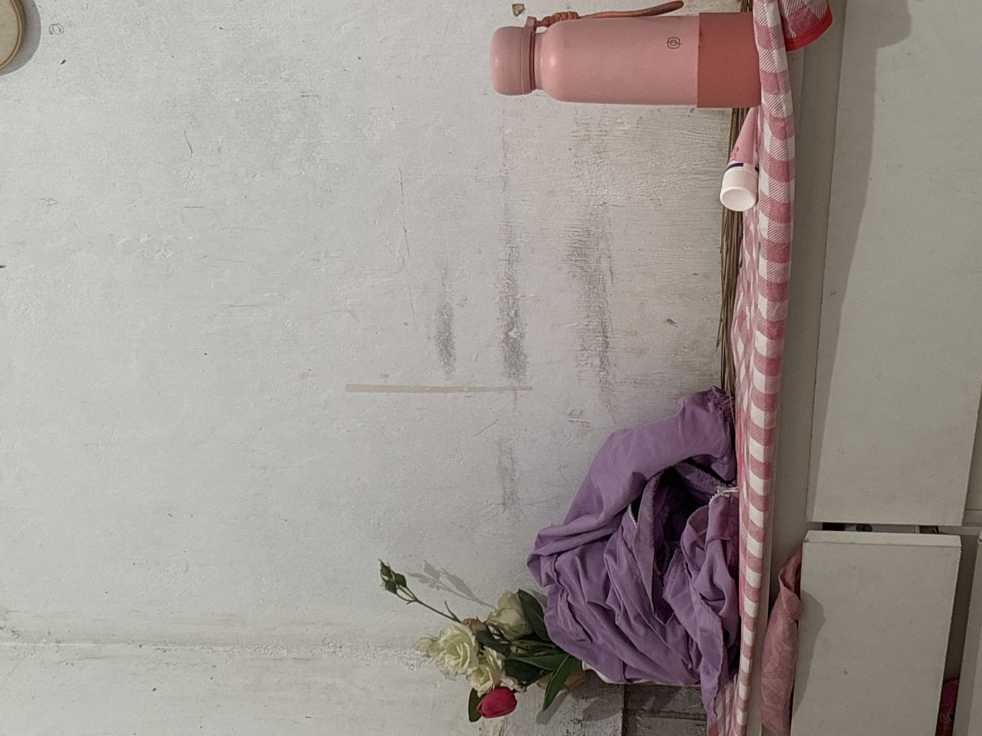 The image depicts a dimly lit corner of a room, likely a bedroom, with a white, textured wall as the backdrop. In the foreground, a white dresser or cabinet with two visible drawers is partially visible. On top of the dresser, a red and white checkered fabric is spread out.
On the left side of the dresser, a vase holds a bouquet of artificial flowers, including white roses and a single red rosebud. A pile of neatly folded purple fabric, possibly clothing or blankets, rests next to the vase.
Towards the right side of the dresser, a pink water bottle with a white cap and a red string attached stands upright. Next to the water bottle, a small tube, possibly a cosmetic or medication, lies horizontally with its cap off.
The overall impression is of a personal space, possibly a bedroom in North Jakarta, Indonesia, with a few decorative and functional items arranged on a surface. The lighting is subdued, casting soft shadows and giving the scene a sense of calm.
