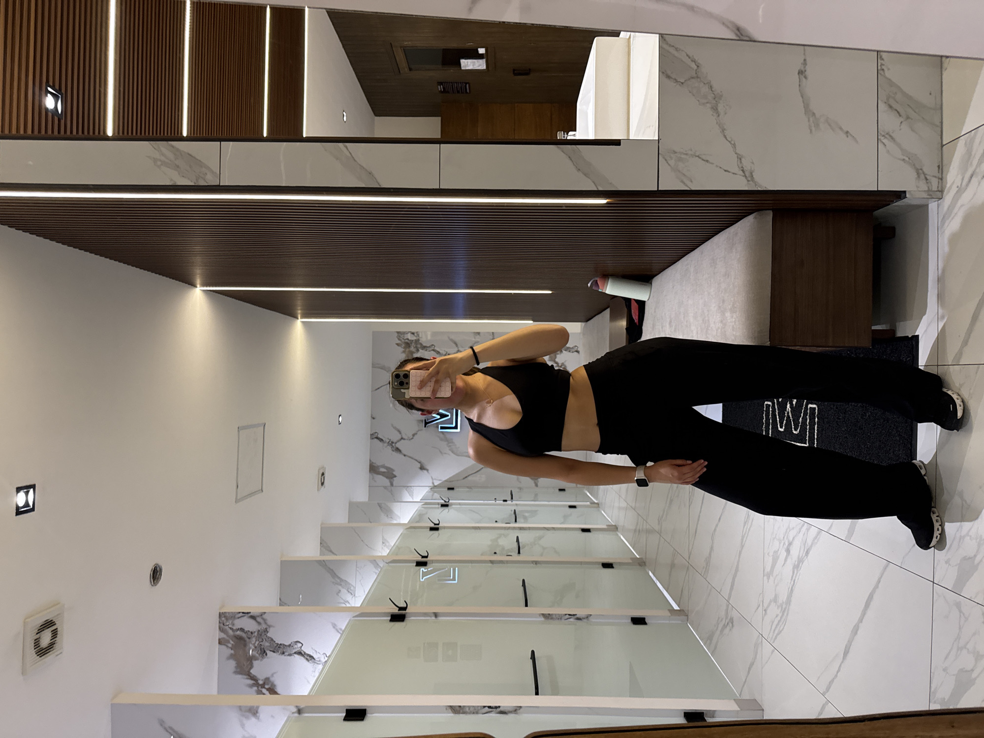 A woman stands in a dressing room, holding a smartphone in her left hand to take a photo of herself in the mirror. She's wearing a black sports bra and black leggings. The dressing room has individual stalls with frosted glass panels. The walls and floor are tiled with white marble-patterned tiles. Above, the ceiling is composed of wooden slats with integrated lighting. The scene is possibly located in a high-end retail space. A rug is visible, bearing a logo. It's likely the location is within Puebla, Mexico.