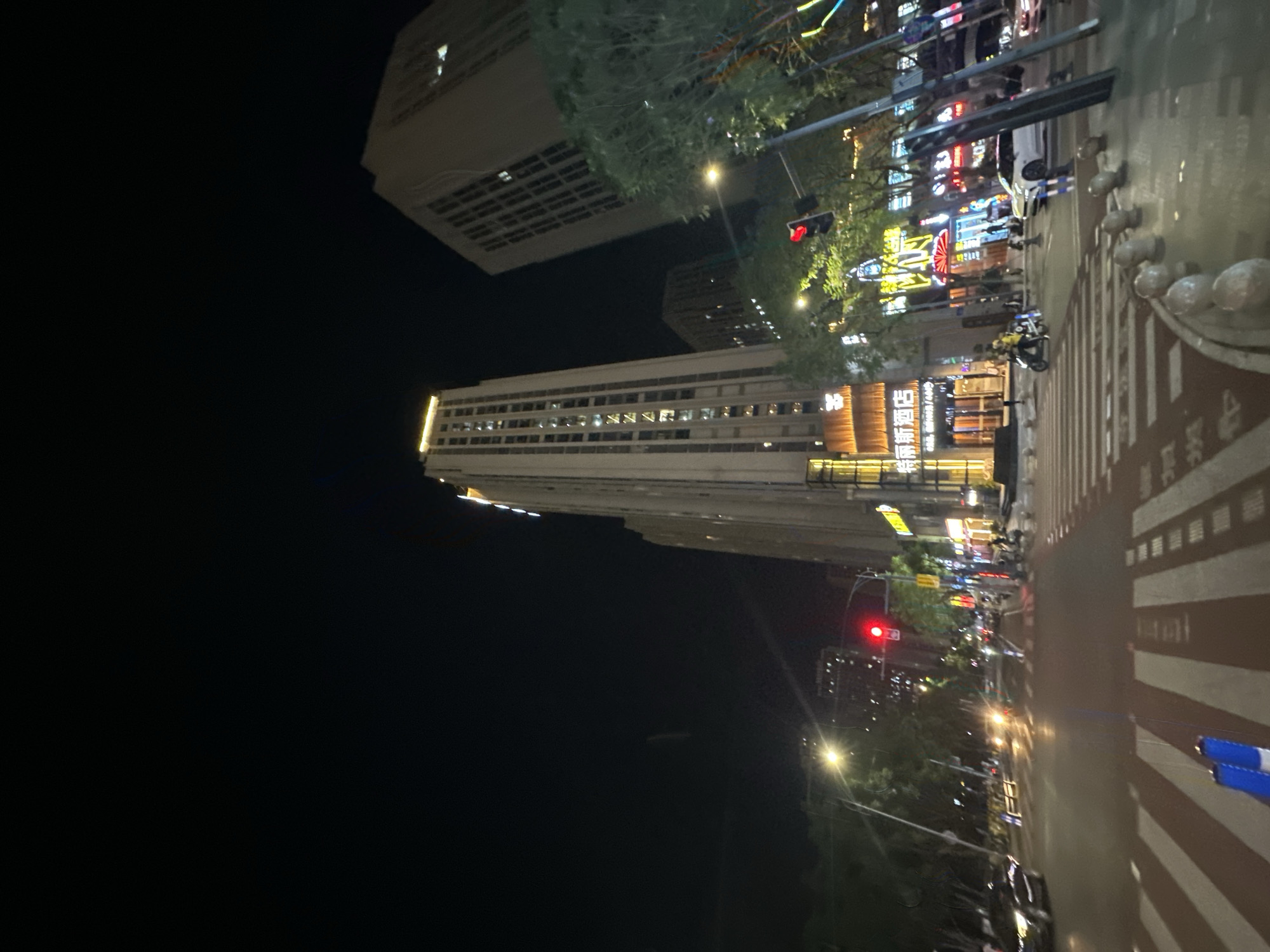 The image shows a nighttime urban scene in Chongqing High-tech Zone, China. Dominating the view are tall buildings lit with rows of windows, interspersed with denser clusters of illuminated commercial signs. Streetlights create a bright zone along the right-hand side, where pedestrian crosswalk markings are visible. A traffic light is illuminated in red. Cars and pedestrians are present, though indistinct. The left side of the image is dominated by a dark night sky.
