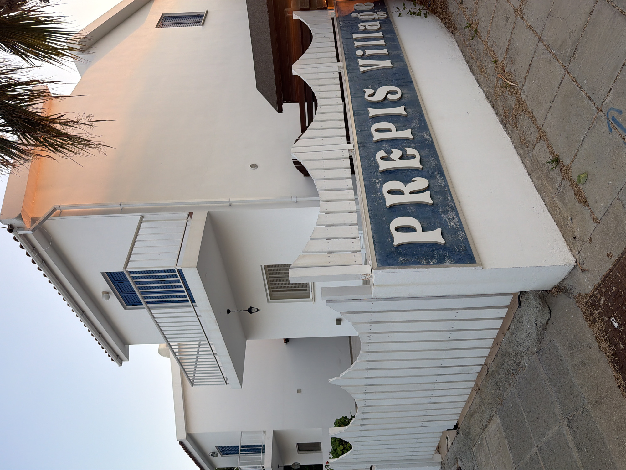 The image shows a white building with blue shutters and a balcony, situated in Pervolia, Cyprus. A white picket fence with a decorative wavy top is in the foreground, and a blue sign with white lettering reads "PREPIS VILLAGE". The building appears to be a house or a complex of houses, possibly a residential or vacation property. The environment seems to be a street or walkway with paved ground. The lighting suggests it is either early morning or late afternoon, with warm light hitting the side of the building and palm fronds visible in the upper right corner.