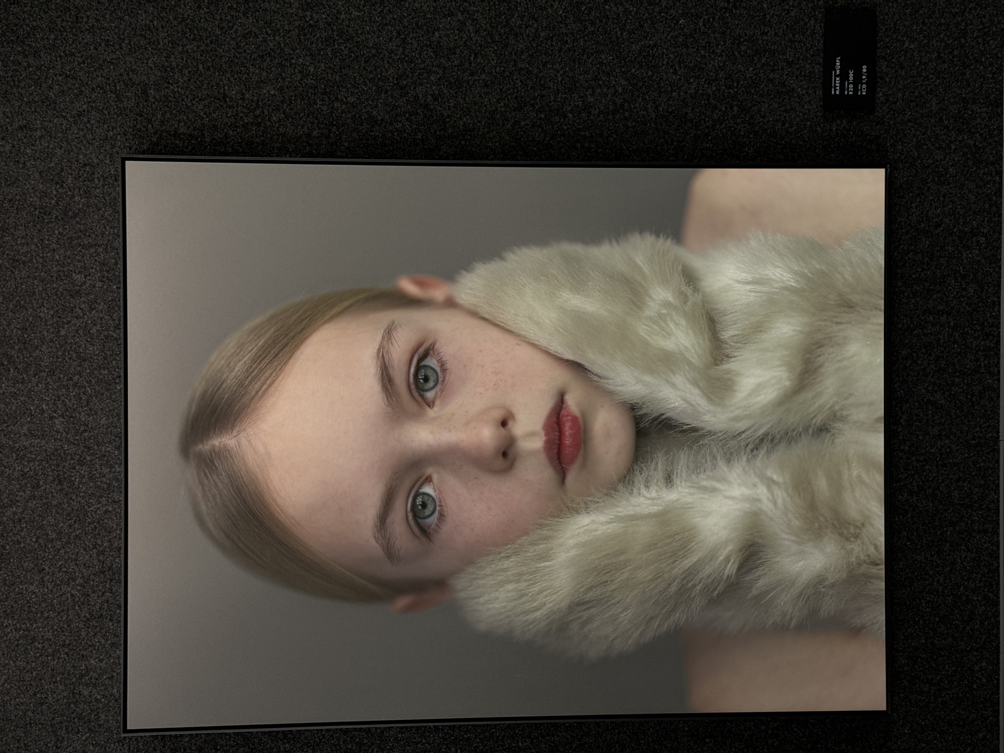 The image shows a framed portrait of a young woman. She has blonde hair neatly styled back and fair skin with freckles. The woman is wearing a pale fur collar. Her eyes are a blue-green color and her lips are red. The portrait is displayed against a dark grey textured background, resembling a wall covering. In the upper right corner is a small black label with white text. The text on the label is "MAREK WÜRFEL", "X2D 100C", and "XCD 1,9/80". It appears the photo was taken at an art exhibition. It is unknown if the photo was taken in Nanshan District, China.