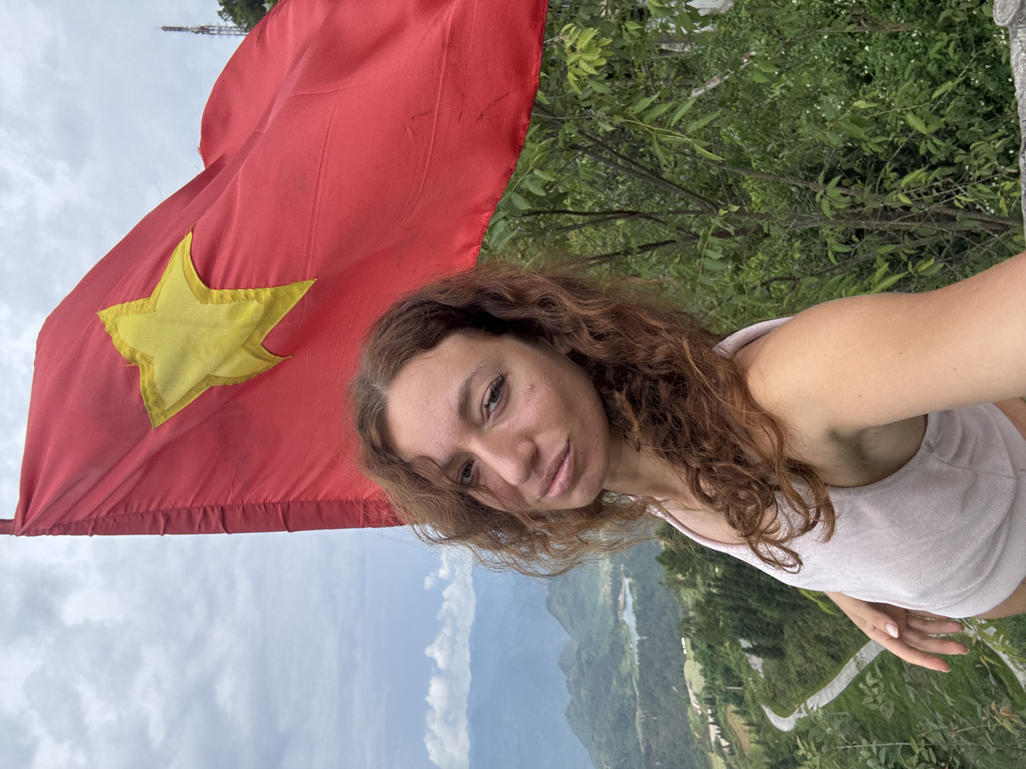 A woman stands in Quản Bạ Commune, Vietnam, posing for a selfie with the flag of Vietnam prominently displayed. The background features a landscape of mountains, trees, and a winding road. The sky is overcast.