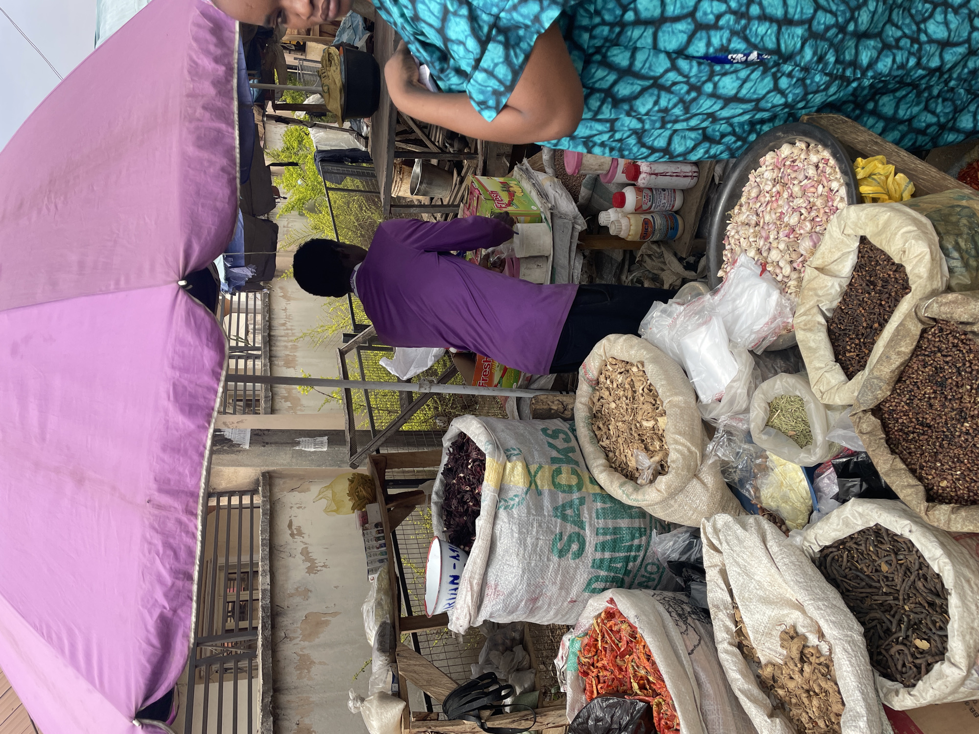 The image shows a market stall in Jos, Nigeria. A man in a purple shirt is interacting with a customer, while another person in a blue top stands nearby. The stall is covered by a pink umbrella, providing shade. Large sacks of various dried goods, including spices, herbs, and possibly grains, are displayed on the ground. Bottles and packages of goods are visible on a raised surface behind the man. The scene appears busy, with other structures and buildings visible in the background, indicative of a bustling marketplace setting.