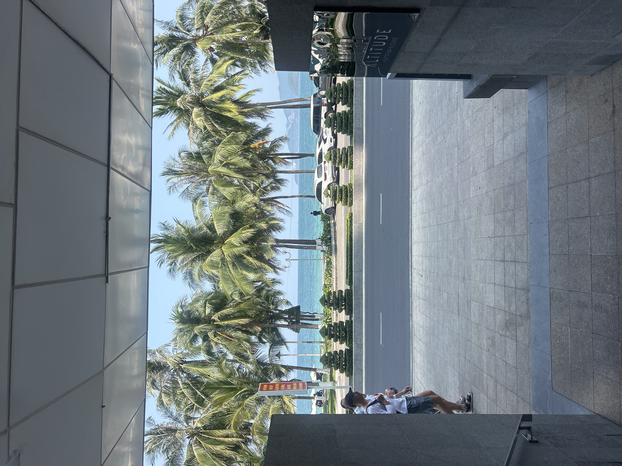 The image depicts a sunlit coastal scene, likely in Nha Trang, Vietnam, viewed from a slightly elevated perspective. On the left, a modern building with reflective, light-grey paneled walls partially mirrors the blue sky and surrounding palm trees.
A wide, paved road with white dashed lines runs horizontally across the middle ground, separating the foreground from the ocean. Several cars, predominantly white, are parked along the roadside, interspersed with neatly trimmed green shrubs. Beyond the road, a vibrant line of tall palm trees with lush green fronds stands between the street and the clear, turquoise ocean. The sea stretches out to the horizon, where distant mountains are visible under a bright blue sky.
In the foreground, on the right, a building with dark, textured grey walls features a black sign that clearly displays "ALTITUDE" with "BISTRO BAR LOUNGE" underneath. On the tiled sidewalk below, two individuals are present. One person is seated or leaning against a wall, wearing a white shirt, dark shorts, and sandals, appearing relaxed with their head tilted upwards. Another person is partially visible behind them.
Further down the street, partially obscured by the palm trees, a red and yellow sign with Vietnamese text is visible, though the specific wording is not legible. The scene suggests a warm, clear day, characteristic of a tropical or subtropical resort area.