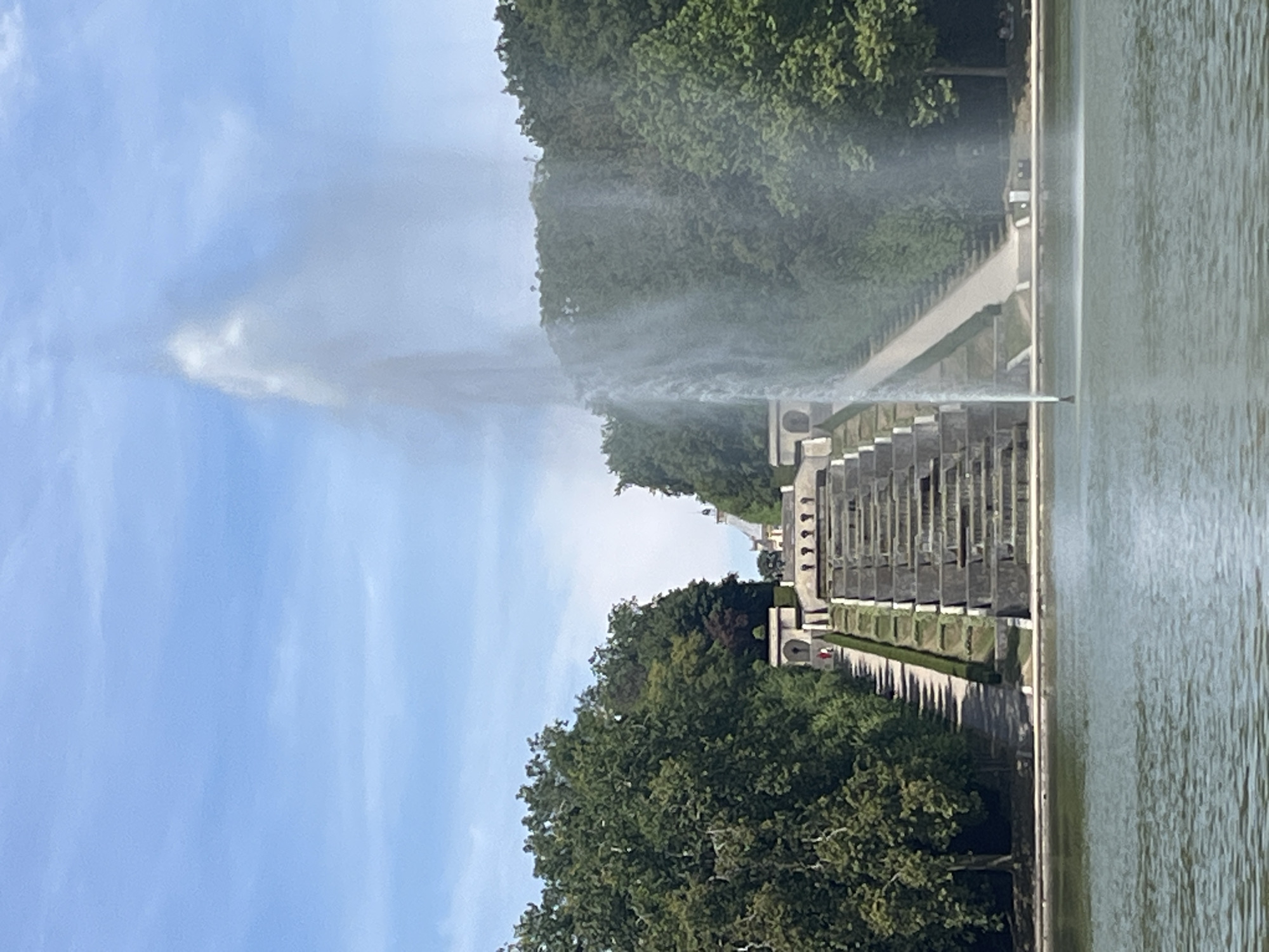 The image shows a grand tiered fountain and cascading water feature, viewed from a body of water in the foreground. The fountain is in the center of the frame, shooting a tall plume of water into the air. Behind the fountain, a long, wide set of stone stairs descends from a building at the top of a hill, flanked by manicured hedges and trees. People are visible on the stairs, appearing as small figures, and a path runs alongside the stairs. The scene is set within a lush green park or garden, with dense trees on either side of the fountain and stairs. The sky above is a clear blue with some wispy clouds. The architectural style of the building at the top suggests a historical or formal garden setting. This appears to be the Parc Sceaux, located in Antony, France, known for its expansive formal gardens and grand fountain.