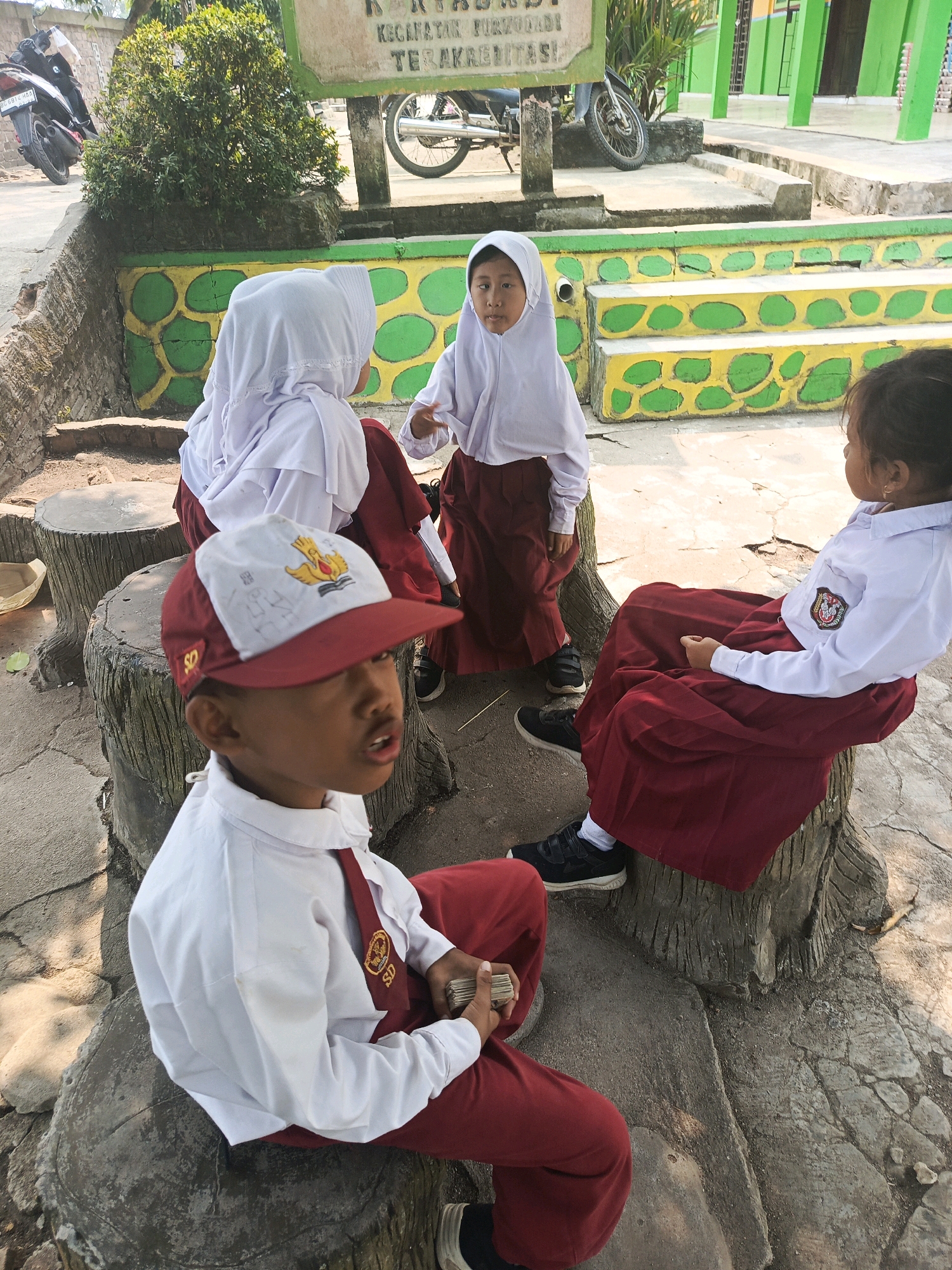 Four children, likely students based on their uniforms, are sitting on tree stumps outdoors. The child in the foreground is a young boy wearing a white shirt, red tie with "SD" insignia, and a red and white baseball cap with an emblem. He is looking upwards with his mouth open. To his left, two other children, a girl wearing a white hijab and a red skirt, and another girl wearing a white hijab and red skirt, are seated. Another girl is seated to the right, also in a white shirt and red skirt, facing away from the camera. The environment appears to be a schoolyard or public area with a sign in the background that has Indonesian text. There are also motorbikes visible in the background. The ground is paved with stone and dirt.