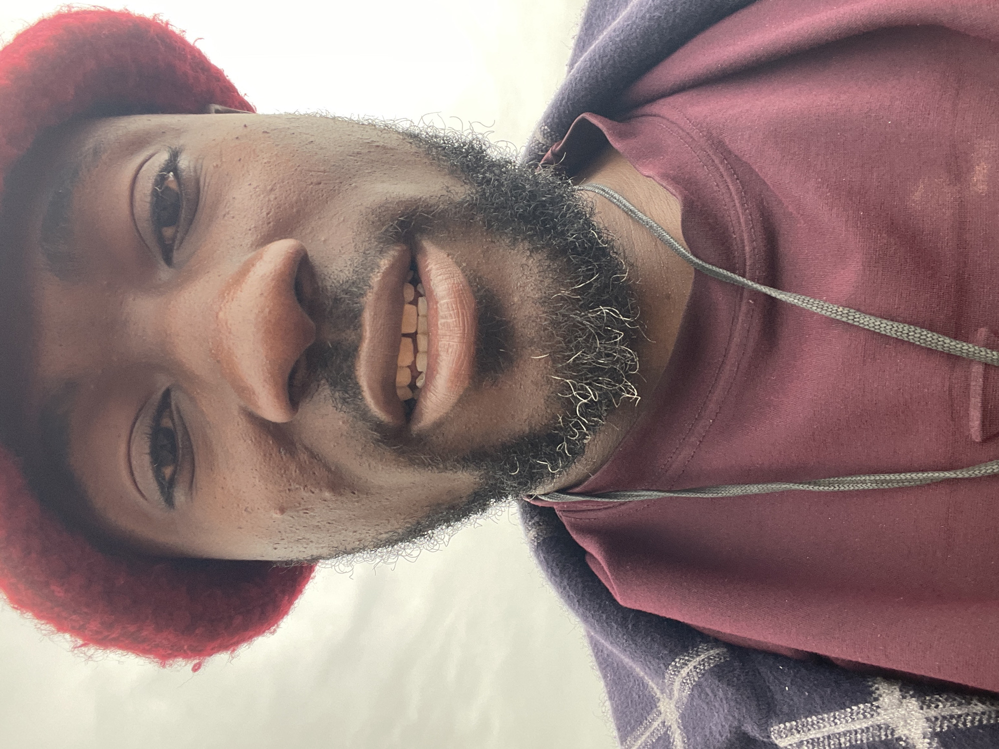 A close-up, eye-level, selfie-style photo shows a Black man wearing a red knitted hat and a burgundy t-shirt layered under a dark plaid jacket. He has a beard with some gray hairs. His mouth is slightly open, revealing his teeth. The background is a hazy, overcast sky. The image appears to be taken outdoors. There is no information about the location in Jos, Nigeria.