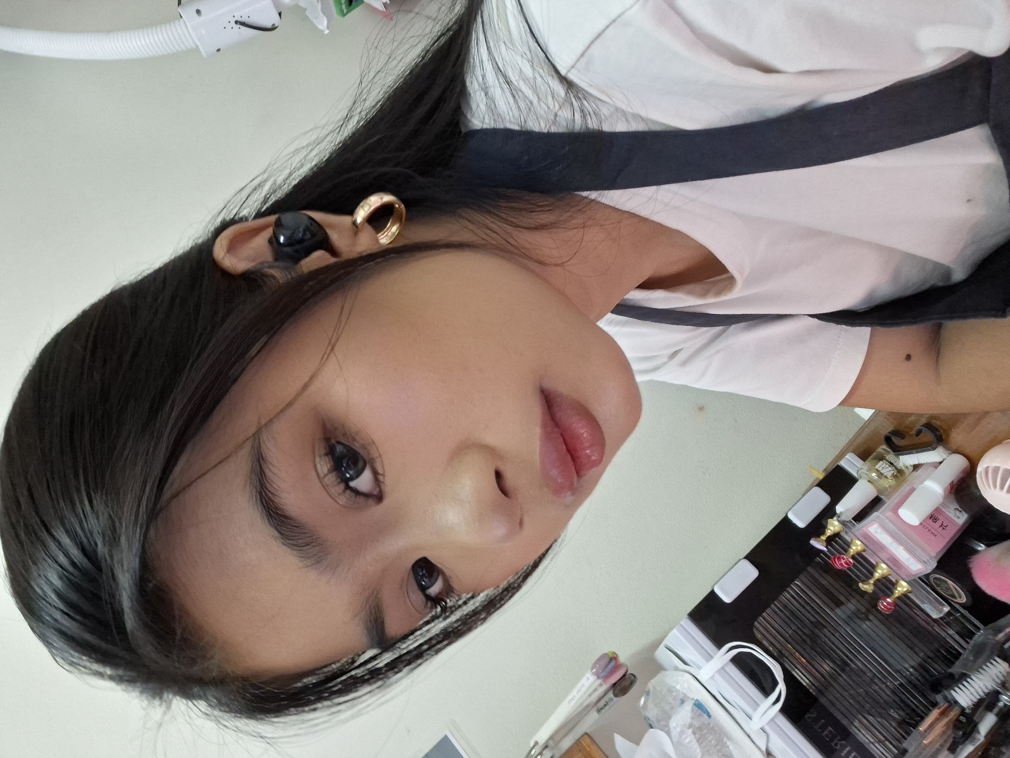 A young woman with dark hair and makeup takes a selfie in an indoor setting that appears to be a nail salon, based on the visible nail art supplies and tools. She is wearing a white t-shirt and a dark apron. She has a gold hoop earring in her left ear and a black wireless earbud in her right ear. The background shows shelves with various beauty products and supplies. The lighting is bright and appears to be artificial. The woman's face is turned towards the camera, and her expression is neutral. The photo is taken from a slightly high angle, looking down at the woman. There is no indication of the city of Kuta, Indonesia.