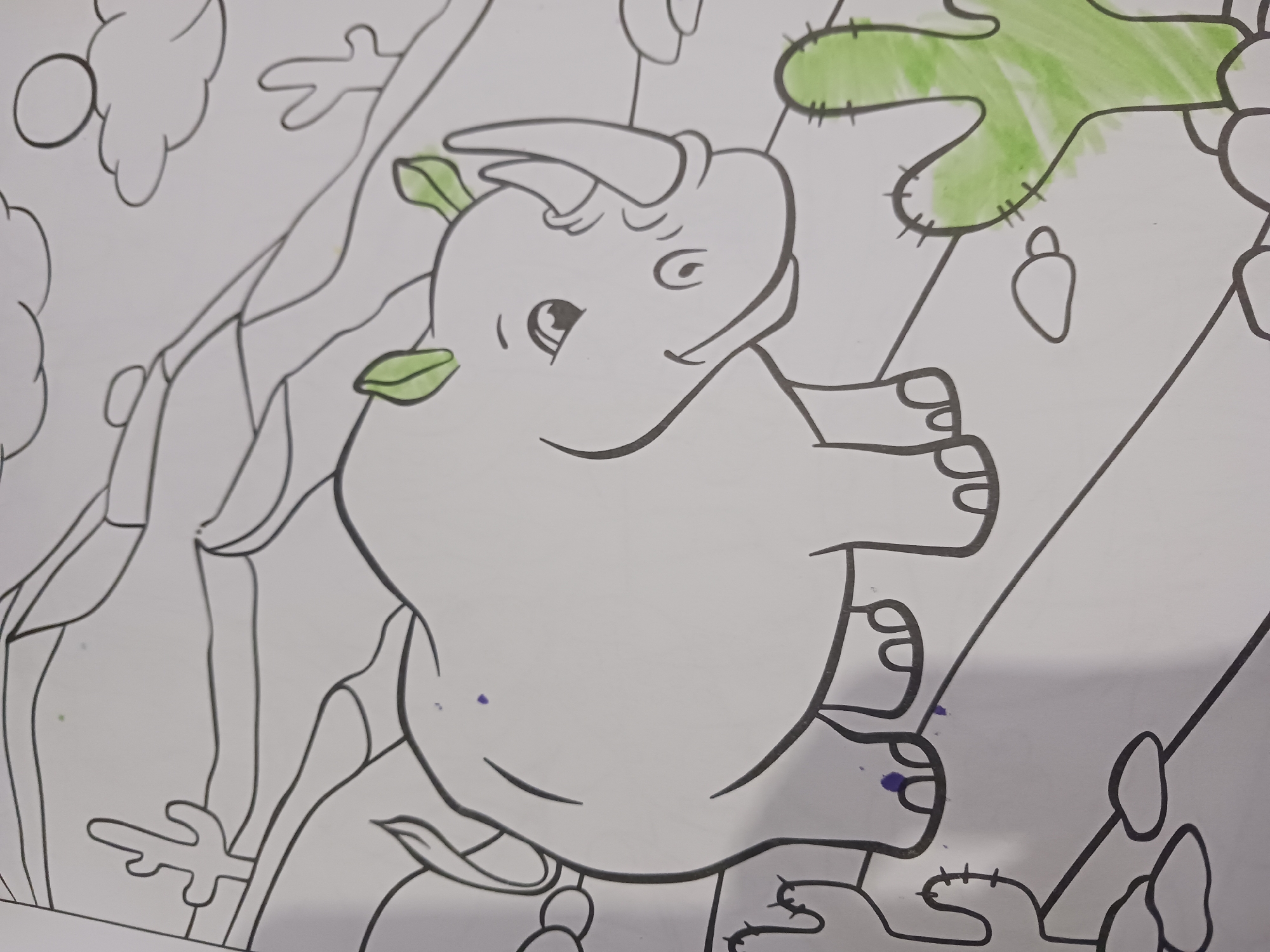 The image is a close-up, slightly angled, eye-level shot of a coloring book page featuring a cartoon rhinoceros in a desert landscape. The rhino is positioned in the center of the frame, facing left with its mouth open in a smile. It has a large horn and green-colored ears. To the left of the rhino, there are two cartoon saguaro cacti, and to the right, a larger saguaro cactus is partially colored green. The background depicts rolling hills, clouds, and a sun. The coloring book lines are black and clearly defined. There are a few small purple smudges on the rhino and one on the ground. The coloring of the cacti is done with a bright green crayon or marker, with some areas showing more intense color than others. The rest of the image is uncolored white paper. The scene is located in Unknown, Unknown.