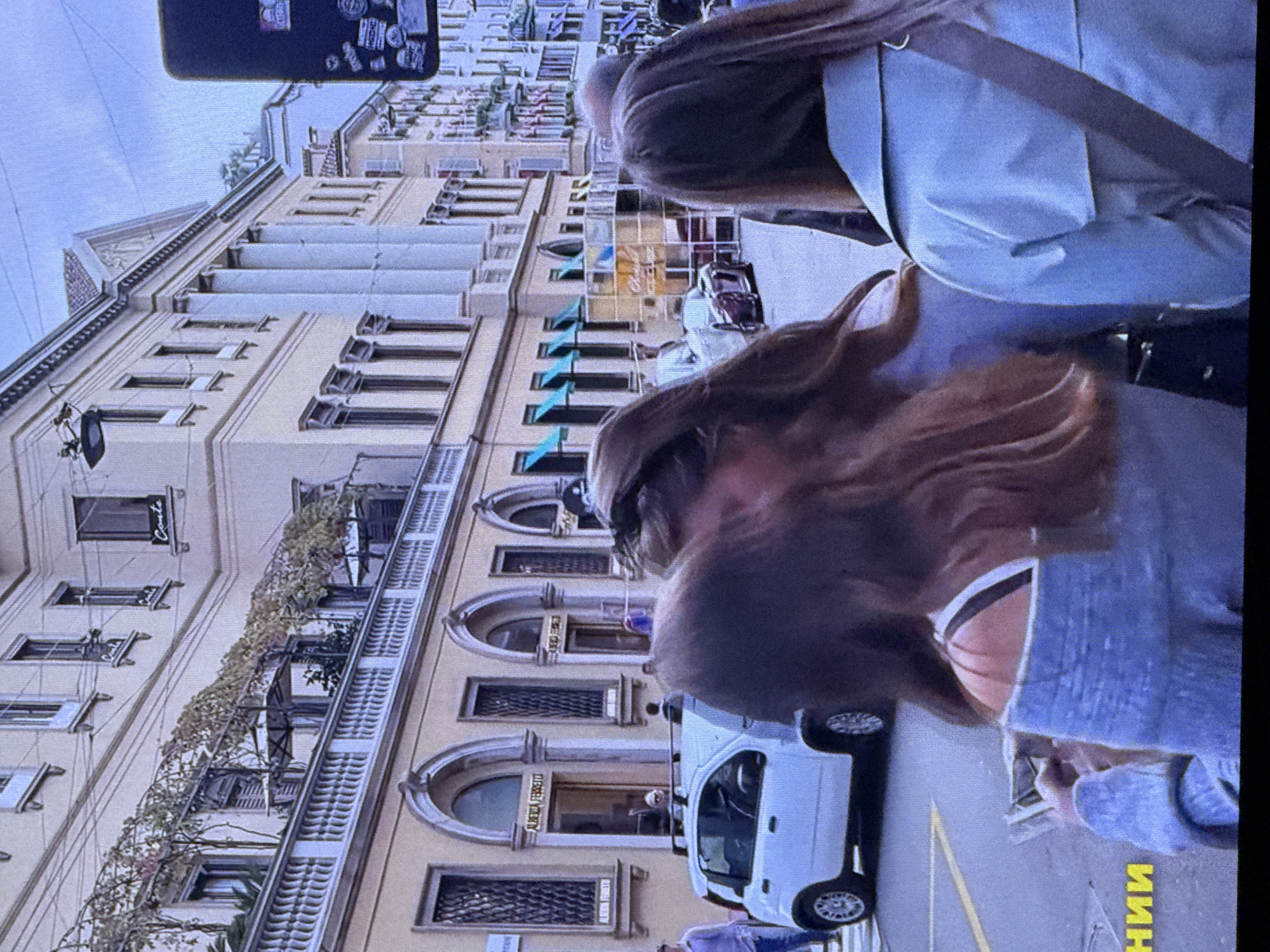 The image displays a street scene in front of a cream-colored building with classical architectural elements, including columns and arched windows. Vines are growing on a balcony on the upper level. A white hatchback car is parked on the street, and several people are visible walking around. In the foreground, the backs of three women's heads and shoulders are prominent. One woman in the center has long, light brown hair. Another to her left has darker hair. The woman on the right wears a light blue jacket with a grey strap across it. The scene appears to be a public area in a city, with the building's facade suggesting a commercial or historical district. There is no indication of the location being Dubai, United Arab Emirates.