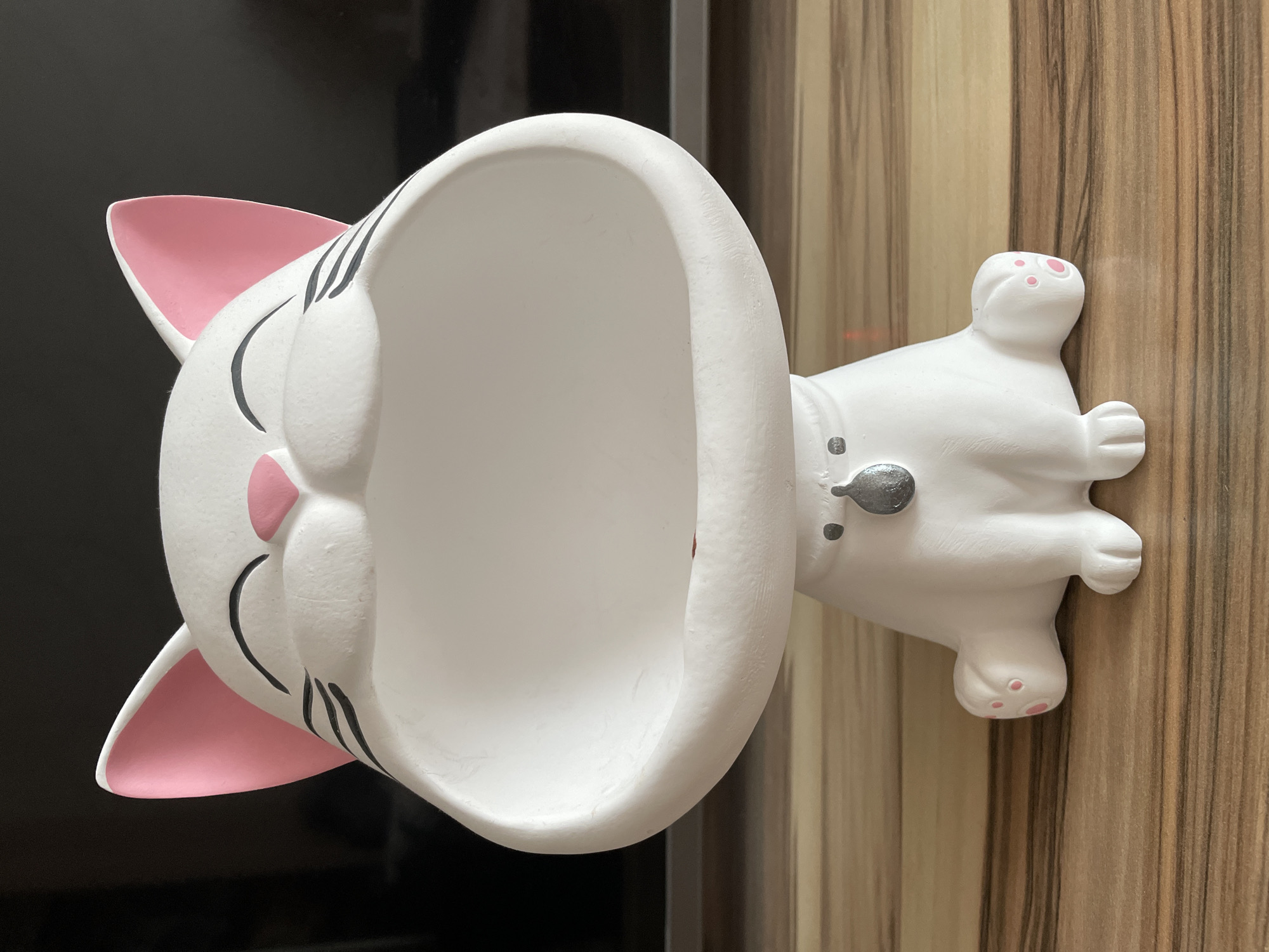 A white ceramic cat-shaped trinket holder sits on a wooden surface. The cat has pink inner ears, pink paw pads, black whiskers, and a large open mouth. It is wearing a silver tag around its neck. The background is dark and out of focus. The image does not contain any information about the city of Nefteyugansk, Russia.
