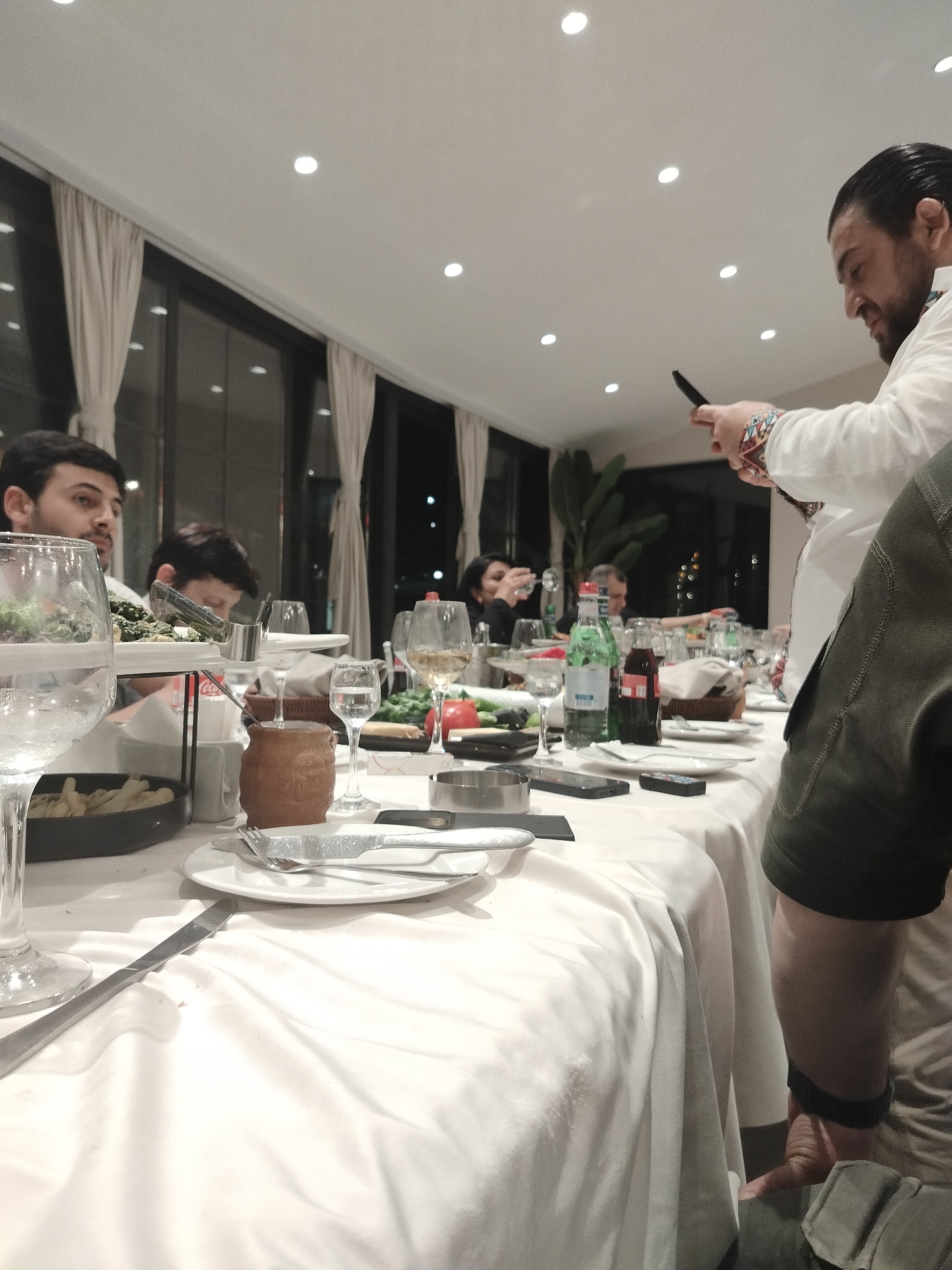 Scene: A gathering in Yerevan, Armenia. A long table covered with a white tablecloth is laden with food, beverages, and tableware. Visible on the table are plates, cutlery, glasses (some filled with water and wine), a small earthen pot, and various dishes.
People: Several people are seated at the table, including two men in the foreground. A man wearing a white shirt with a patterned band on the sleeve is standing and holding a smartphone. A person in a green jacket is visible at the edge of the frame. Other individuals are seated further down the table, appearing to be engaged in conversation.
Objects: The table is furnished with standard dining items like plates, forks, knives, and glassware. Bottles of water and Coca-Cola are present. A smartphone is held by the standing man.
Actions: The man holding the smartphone appears to be taking a photo or video. The seated people seem to be enjoying a meal or social gathering.
Environment: The setting is indoors, likely in a restaurant or dining room. The room is well-lit by ceiling lights, and large windows are visible in the background with dark curtains. A potted plant is also visible in the background.