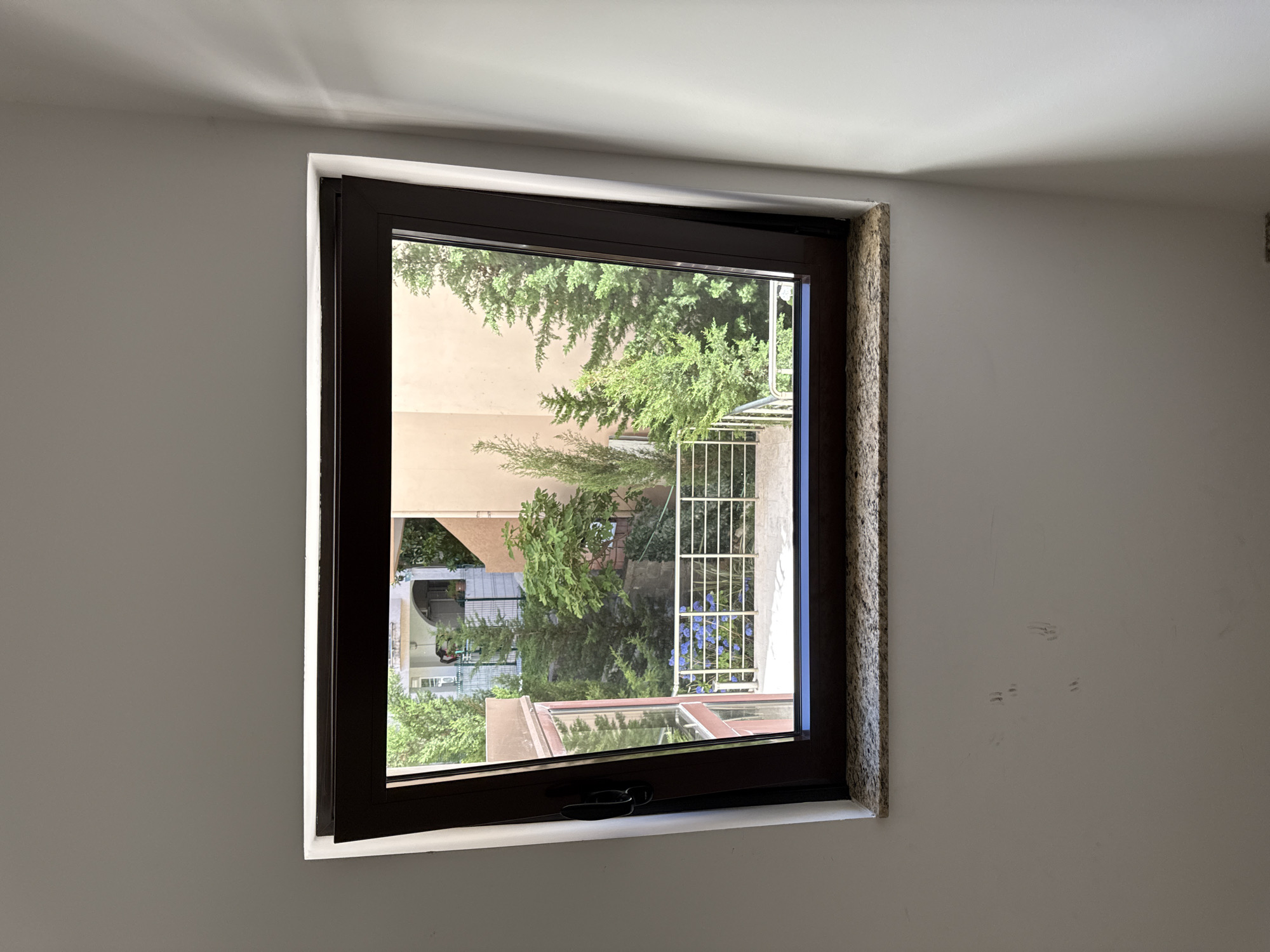 The image depicts a dark-framed window viewed from the interior of a room with white walls. The window is open at the top, allowing a view of an exterior courtyard. The courtyard features a metal railing, a paved area, and an abundance of green trees and some blue flowers. In the background, there are buildings with balconies and windows. The windowsill is made of granite. There are some faint scuff marks on the interior wall below the window. The lighting suggests it is daytime, with sunlight casting shadows on the right wall. The scene is consistent with a residential building in Budva, Montenegro.