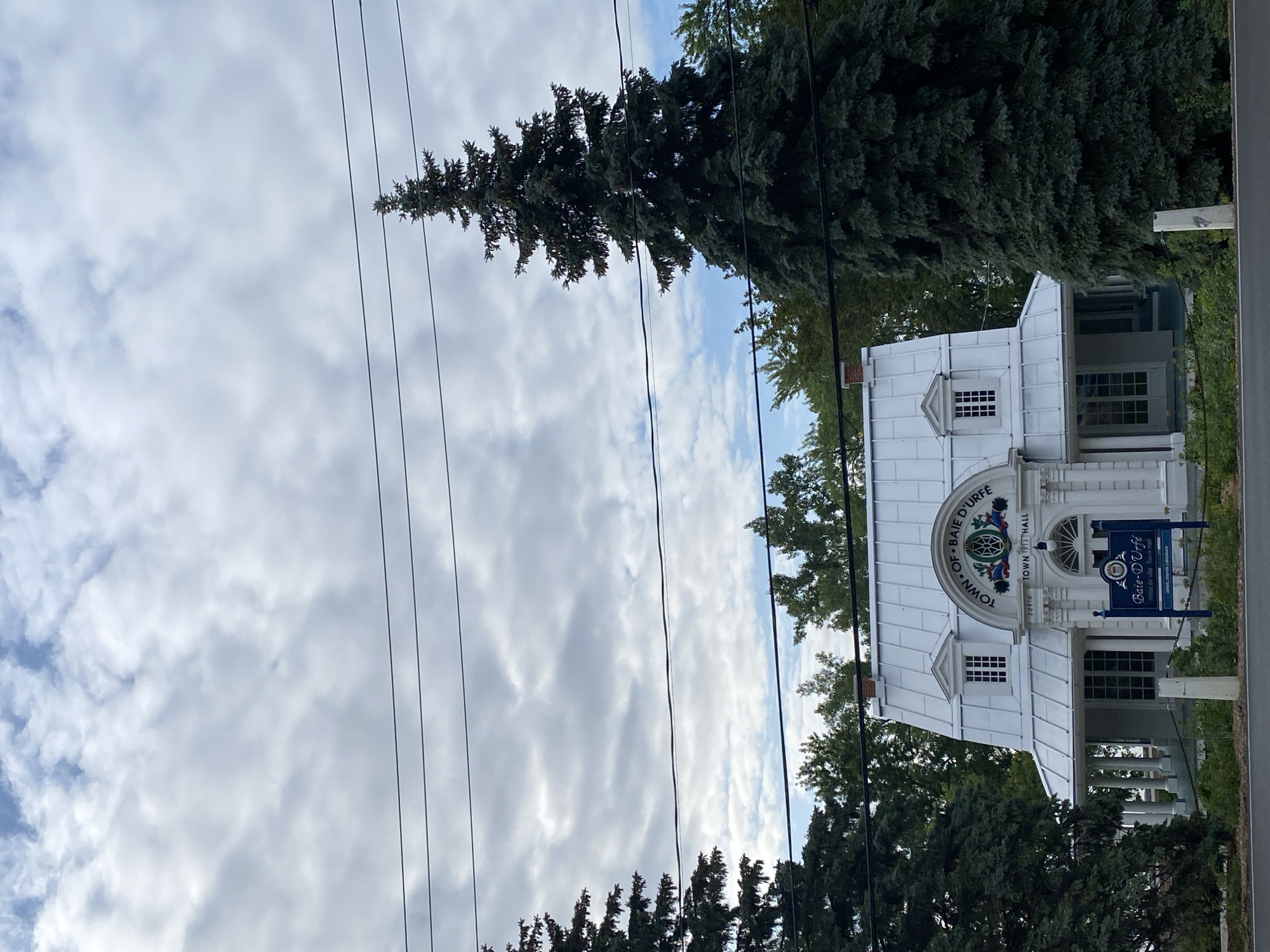 This image depicts the Town Hall of Baie-D'Urfé, Canada. The building, with its white metal roof and classical architectural elements like columns and arched windows, appears to be a historic structure, as indicated by the text "TOWN 1913 HALL" visible on its facade. The building is surrounded by lush green trees, and power lines are prominent in the foreground, crossing the frame. The sky is filled with a mix of white and grey clouds, suggesting an overcast or partly cloudy day. The overall scene is serene and well-maintained, conveying a sense of civic pride and historical significance. The visible text further confirms the location as "TOWN OF BAIE D'URFE" and labels the building as "Hotel de ville Town Hall."