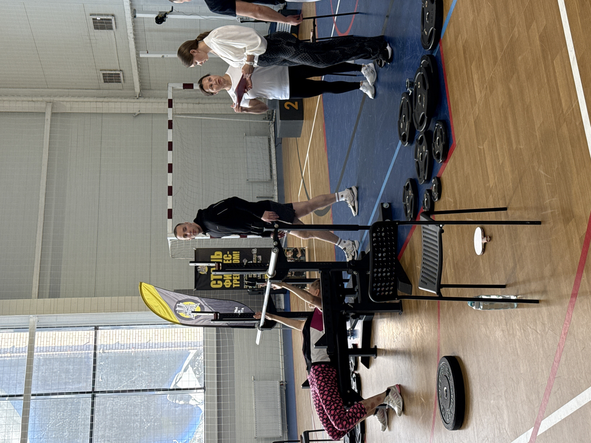 Here is a detailed description of the image:
**Scene:** The image appears to be taken inside a gymnasium in Votkinsk, Russia, with a blue and brown floor and white walls.
**People:** There are several people in the image:
- A woman in pink leggings is using weight training equipment.
- Two women stand nearby, observing and documenting on a clipboard, suggesting they may be coaches or judges.
- A man stands behind the weight training equipment, possibly providing support or monitoring the activity.
**Objects:**
- Weight training equipment is the main focus, including what appears to be a machine for upper body strength training.
- Various weight plates are stacked near the equipment.
- A banner with text in Russian is visible in the background, indicating the gym or competition name and sponsorship.
- A soccer goal is in the background, signifying the gym's multifunctional design.
**Actions:**
- The woman is actively engaged in weight training.
- The two women observe and record, implying an evaluation or monitoring role.
- The man is watching and possibly assisting.
**Environment:** The gymnasium has an indoor sports facility layout, with weight training equipment set up on one side. Large windows provide natural light.