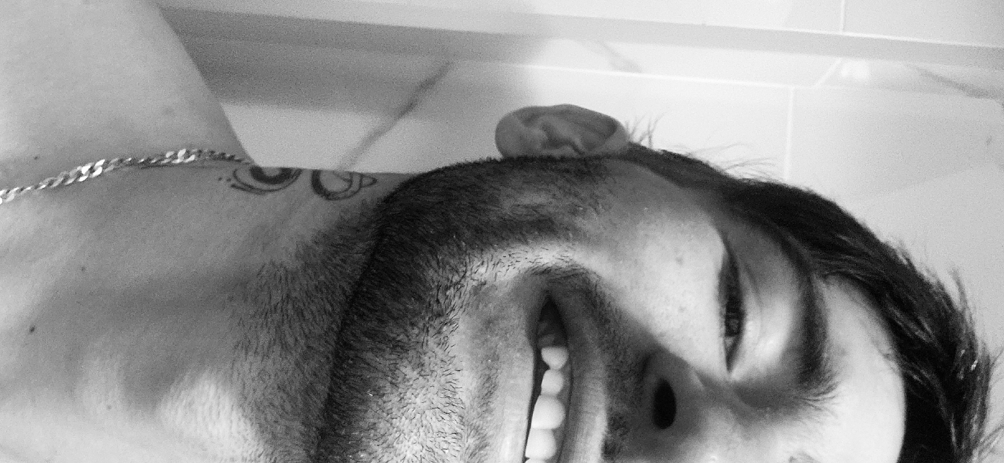 A black and white, close-up photograph captures a section of an adult male's upper body and face, oriented horizontally due to a counter-clockwise rotation. The subject has dark hair and visible stubble or a short beard on his face.
His face is partially visible, featuring one eye, part of his nose, and an open mouth revealing a row of white, even teeth, indicating a smile or laugh. A silver chain necklace is worn around his neck, and a tattoo with abstract or symbolic elements, possibly resembling an eye, is visible on his shoulder/neck area.
The background appears to be a light-colored, possibly tiled wall, suggesting an indoor setting. The expression on the man's face conveys happiness or amusement. There is no visible text in the image. While the location context is Plovdiv, Bulgaria, this is not visually evident from the photograph itself.
