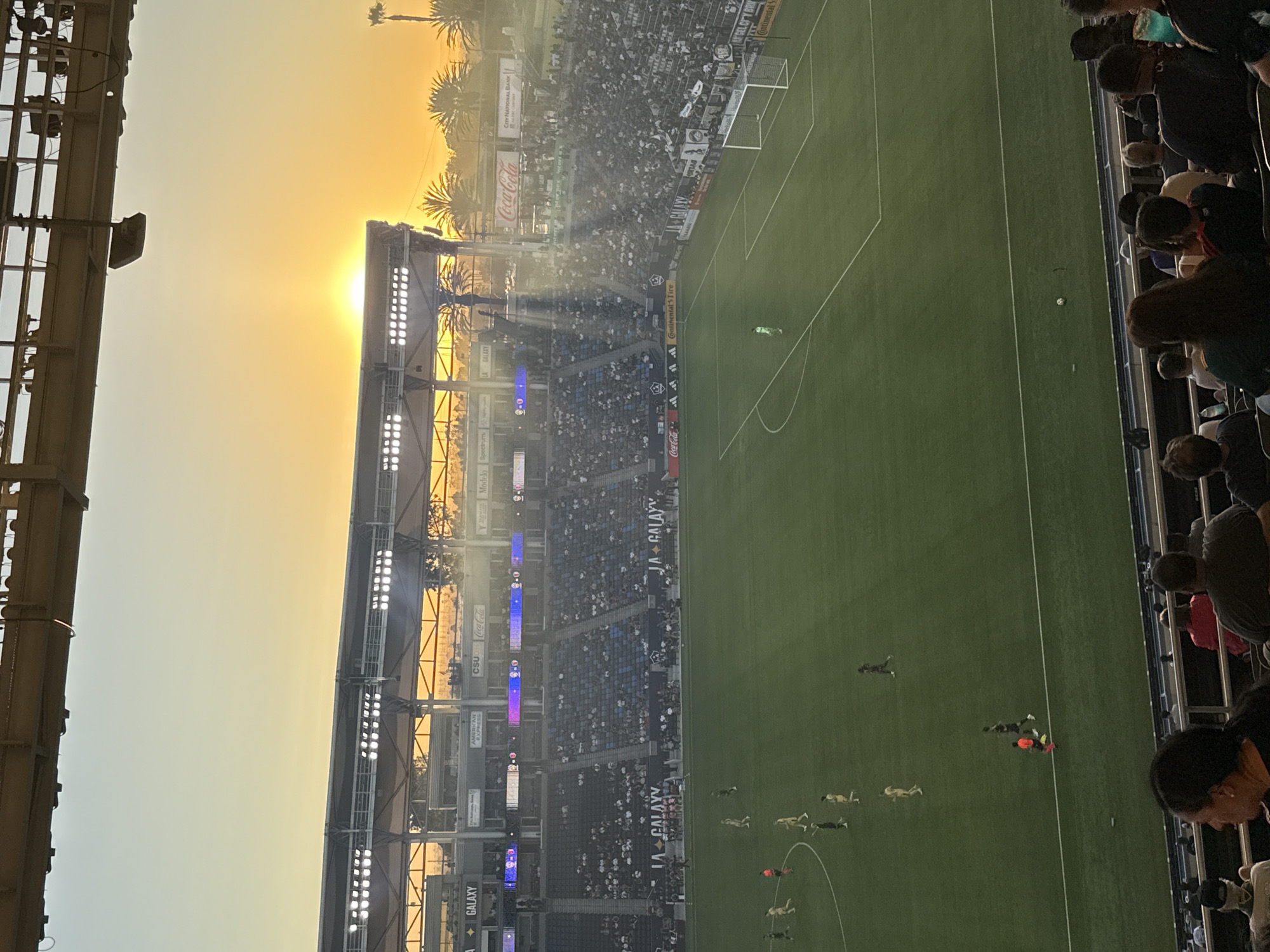 The image captures a soccer match at Dignity Health Sports Park in Carson, California, USA, during sunset. The sun is low on the horizon, casting a warm orange glow across the sky and illuminating the stadium with dramatic rays of light. Spectators are seated in the stands, some visible in the foreground and others filling the tiered seating areas around the field. Players in uniform are on the green soccer pitch, engaged in the game. The stadium infrastructure, including lights and advertising boards, is visible. The overall scene depicts a lively sporting event in an outdoor stadium setting at dusk.