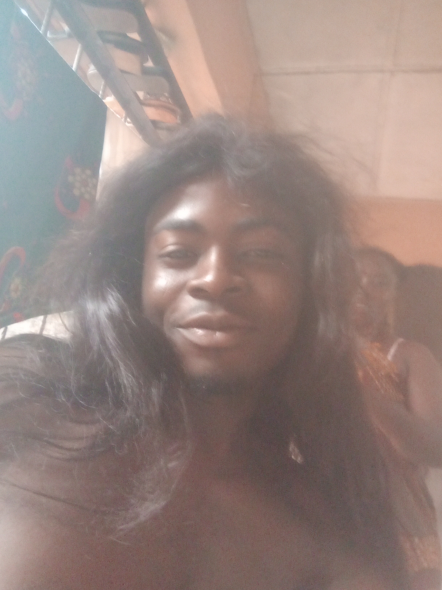 A selfie taken indoors shows a Black man with long, dark hair that appears somewhat disheveled. He is smiling slightly, looking directly at the camera. Behind him, another person, possibly a woman, is partially visible. The background is indistinct, but suggests an interior setting. Based on the location mentioned in the prompt, this scene is set in Ogidi, Nigeria.