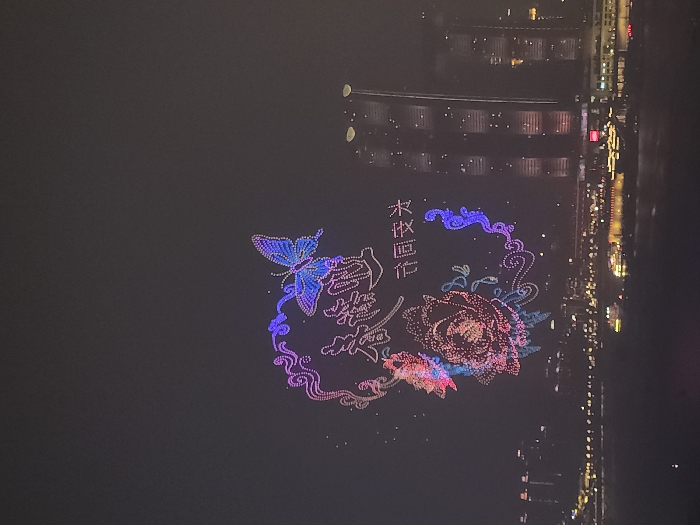 This nighttime image depicts a drone light show over the city of 江北城街道, China, positioned above a major river and urban skyline. The central focus is a large, elaborate drone formation rendered in vibrant blue, purple, pink, and red lights. The design features a prominent blue and purple butterfly at the top right, a large red and pink blooming flower (possibly a peony or rose) centrally, and intricate swirling patterns framing these elements. Two lines of Chinese characters are also formed by drones: "喜迎春光" (Joyfully welcome the spring light) at the top, and "奋同发力" (Strive together to exert force) below it. In the background, numerous multi-story buildings, including skyscrapers, are illuminated across the cityscape, particularly prominent on the right side of the frame. A wide body of water, likely a river, occupies the lower third of the image, upon which several illuminated boats are visible. The sky is dark, indicating a nighttime setting, with no discernable human figures visible.