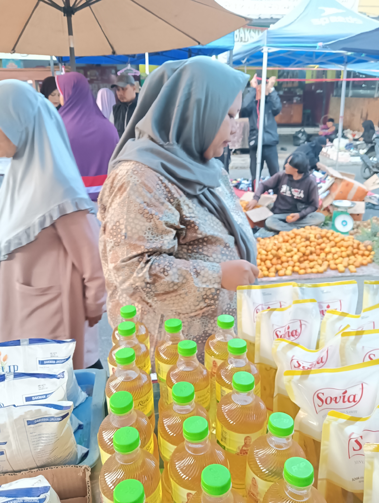 In Katapang, Indonesia, at an outdoor market, a woman in a gray hijab and a patterned brown tunic stands facing right, appearing to examine a display of items. In front of her are rows of clear plastic bottles filled with yellow liquid, each topped with a green cap. Behind her, other women in hijabs are partially visible. To the right, a large pile of small, round, orange fruits is spread out on the ground, with a green and white scale nearby. Other market stalls and tents are visible in the background, along with other people and various goods, suggesting a bustling commercial environment.