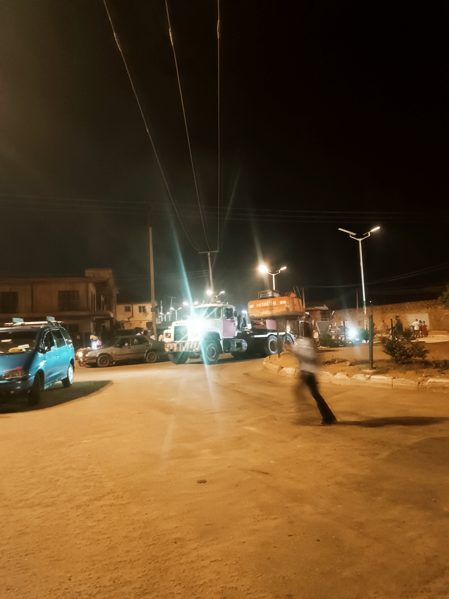 The image depicts a night scene at a roundabout in Bukuru, Nigeria. Streetlights illuminate the area. In the foreground, a blue vehicle is parked to the left. A blurred figure is walking across the road. Further ahead, a truck is carrying a Hitachi excavator. Several people are visible on the right side of the roundabout. The background features buildings and power lines stretching across the sky. The road surface appears to be unpaved.
