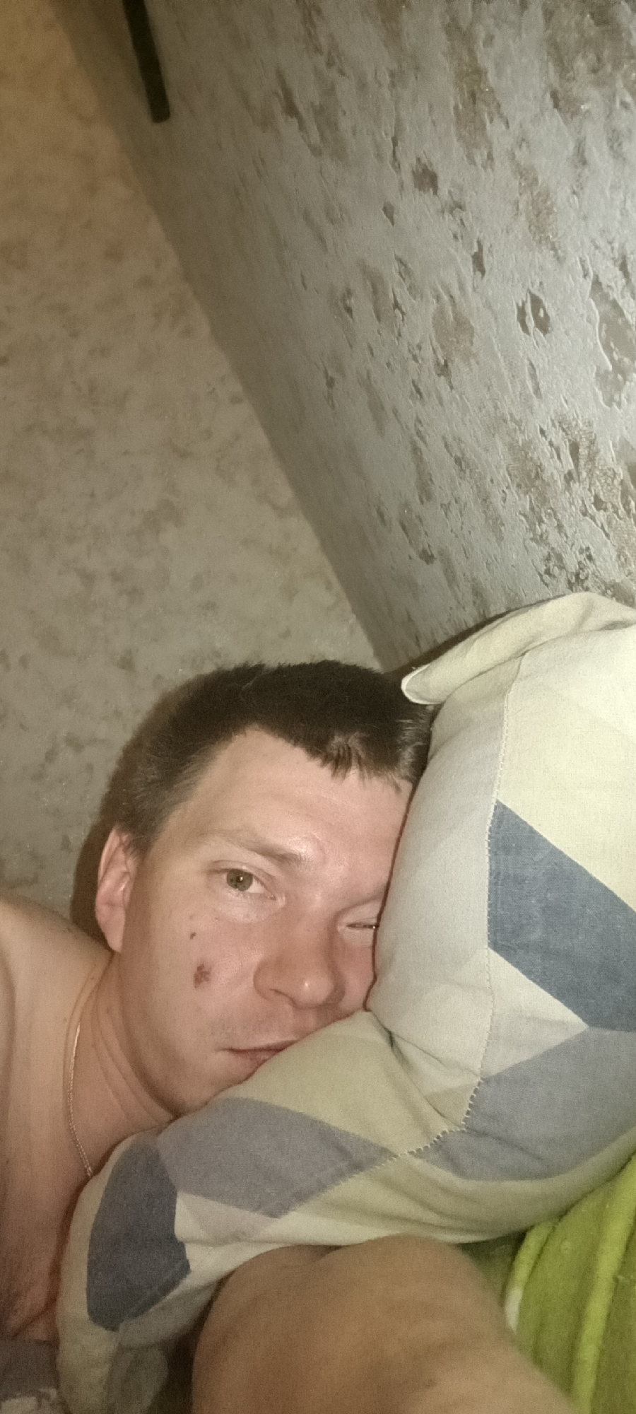 An indoor, eye-level shot depicts a male subject lying on a bed with his head resting on a pillow in the city of Марковский, Russia. The male, with short dark hair, is oriented horizontally, facing the camera. His left eye (viewer's right) is open, while his right eye (viewer's left) is partially closed. A dark, irregular mark is visible on his left cheek. He appears shirtless, revealing a gold-colored chain around his neck. He is partially covered by what appears to be a lime-green sheet or blanket, and his head rests on a pillow with a patterned cover featuring light green, cream, and blue geometric shapes. The background consists of a textured, light brown/beige wall with an uneven surface, and a thin, dark vertical object is visible in the upper left corner of the frame.