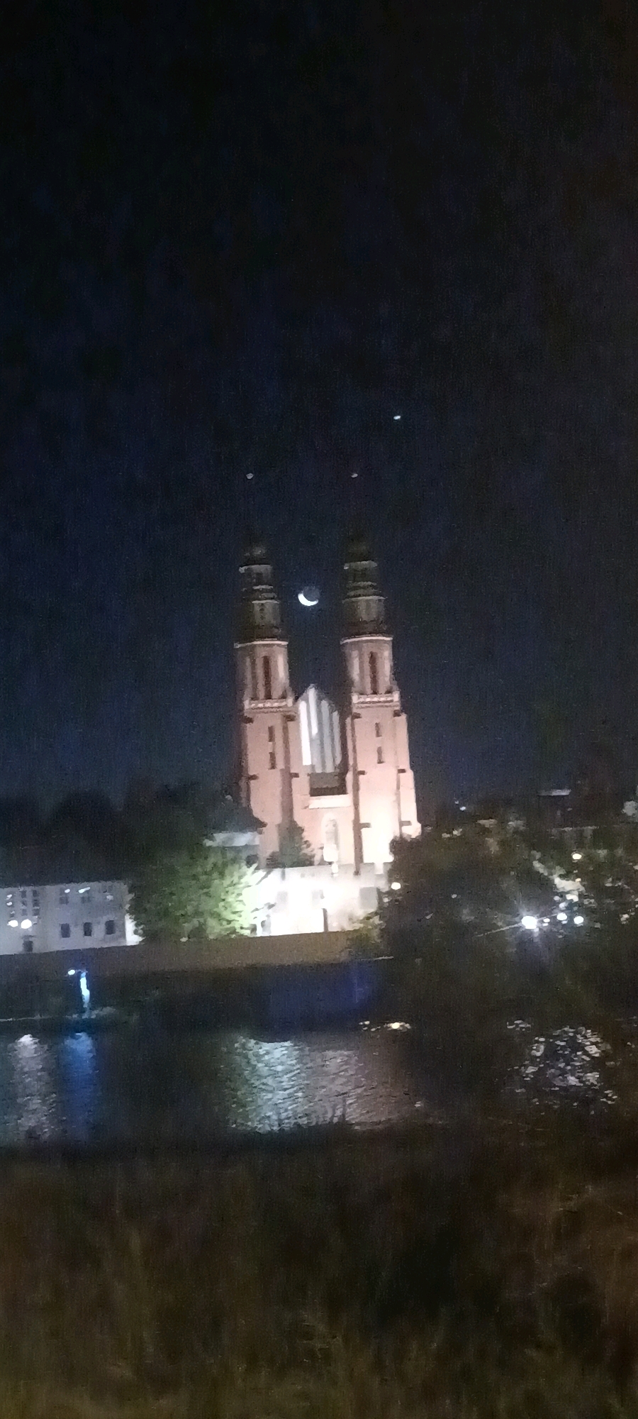 The image captures a nighttime scene in Opole, Poland, featuring the historic Cathedral of the Holy Cross and St. Hedwig. The cathedral's twin towers dominate the center of the frame, illuminated and silhouetted against a dark, starry sky. A crescent moon is visible between the towers, alongside two faint celestial bodies. In the foreground, a body of water, likely the Oder River, reflects the lights of the city and the moonlit sky. The opposite bank of the river shows a line of buildings, also illuminated at night, and a lush green embankment. The overall atmosphere is serene and evocative of a peaceful evening in the city.