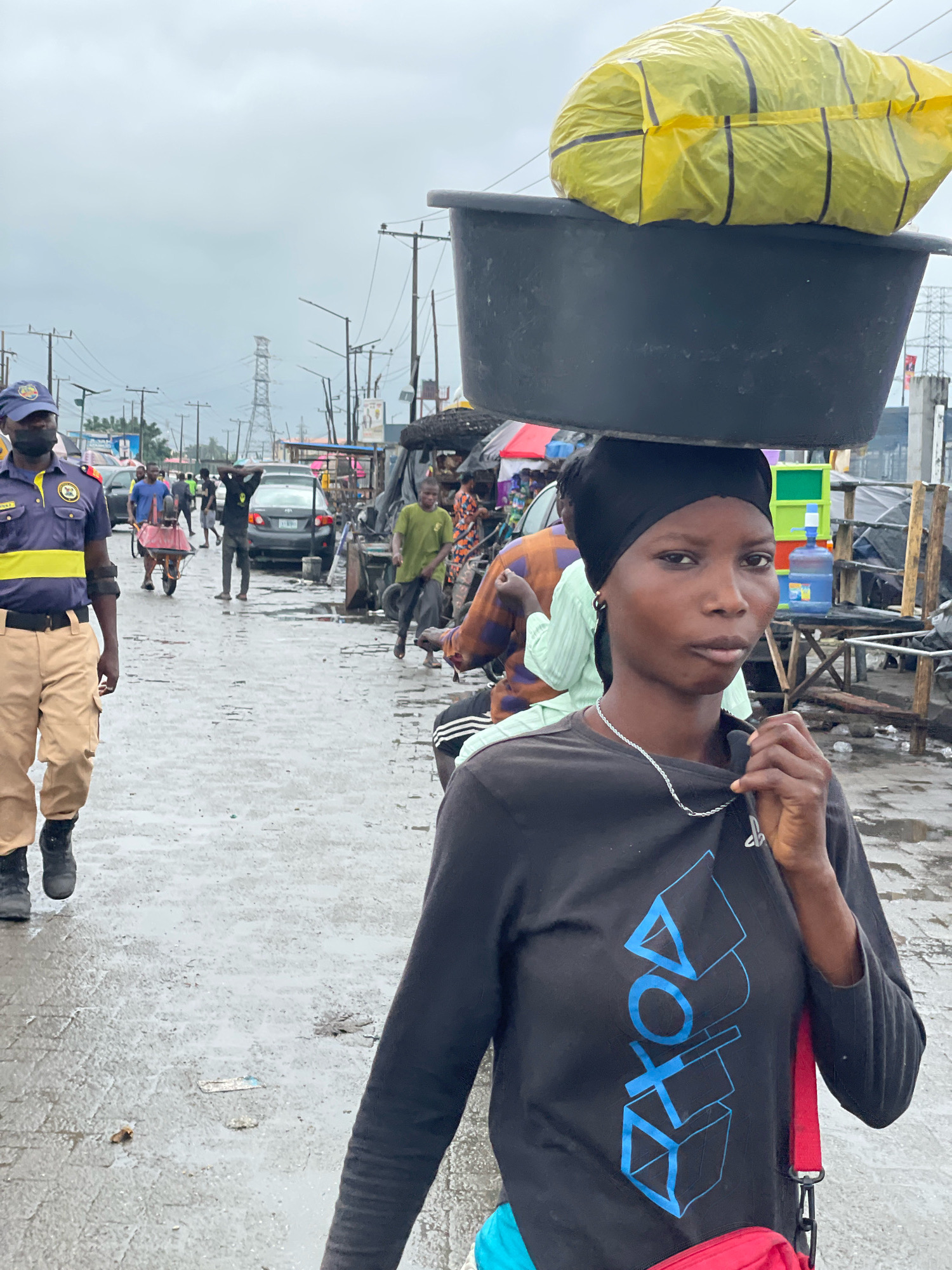 In Ajah, Nigeria, a woman walks carrying a large black basin filled with a yellow object balanced on her head. She wears a black headscarf and a black long-sleeved shirt with blue PlayStation symbols. The ground is wet and appears to be a street or market area, with various people and vehicles visible in the background. A uniformed security officer is also present, walking to the left of the woman. The atmosphere is overcast.