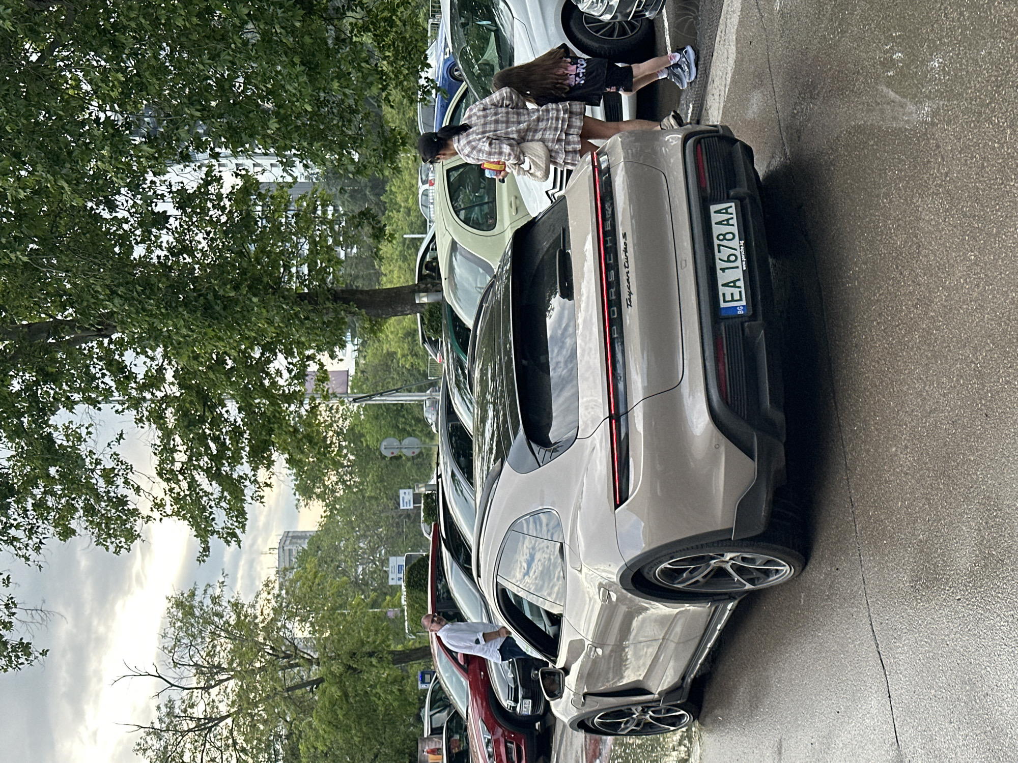 The image captures a row of parked cars on a street in Varna, Bulgaria, characterized by a gray Porsche Taycan Turbo S parked at the end. A woman in a plaid dress stands near the Porsche, possibly interacting with it. There are a few more people near the other cars. The license plate of the Porsche reads "EA 1678 AA", indicating a registration in Bulgaria. Green foliage and trees are visible, along with a building in the background. The street surface is wet, suggesting recent rain.