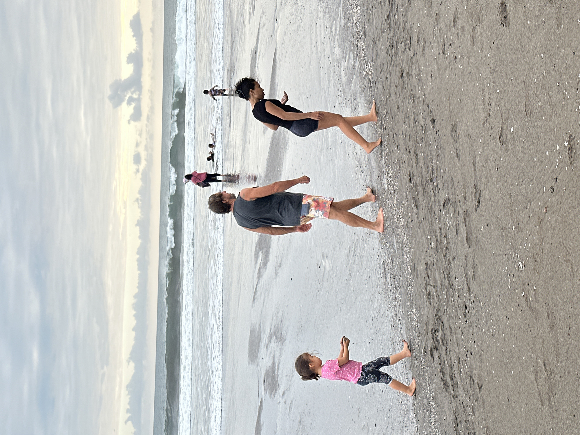 In the city of North Kuta, Indonesia, a family walks along a sandy beach under a cloudy sky. The waves are breaking in the distance, and several other people are visible in the ocean. In the foreground, a young girl in a pink shirt and patterned leggings walks to the left, holding a handful of sand. To her right, a man wearing a dark tank top and colorful patterned shorts walks in the same direction. Further to the right, a woman in a black top and dark shorts walks with her head bowed, looking at her phone. The wet sand is covered with small shells.