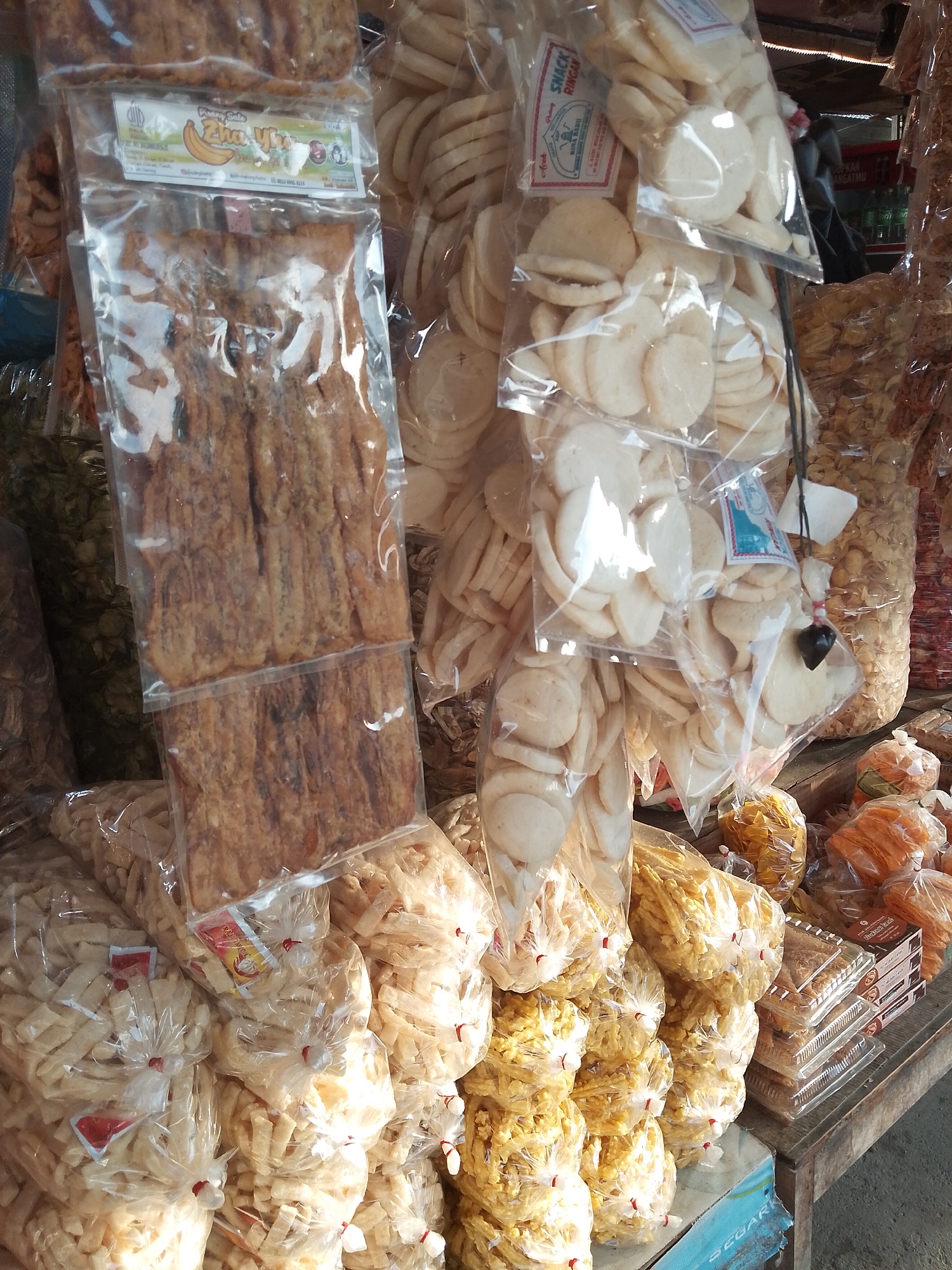 The image captures a stall selling a variety of packaged snacks, likely at a street market in Sawang, Indonesia. In the foreground, several large plastic bags are filled with light-colored, crunchy snacks, possibly crackers or puffed rice. These bags are tied shut with red string. Behind these, a prominent package contains what appears to be dried squid, presented in strips. To the right, more packaged snacks are visible, including some circular, pale-colored crackers and other items in orange and yellow hues. Some of these snacks are neatly stacked in clear plastic containers. The stall itself appears to be made of wood, and the overall setting suggests an outdoor market environment. The lighting indicates it is daytime. There are no people clearly visible in the frame.