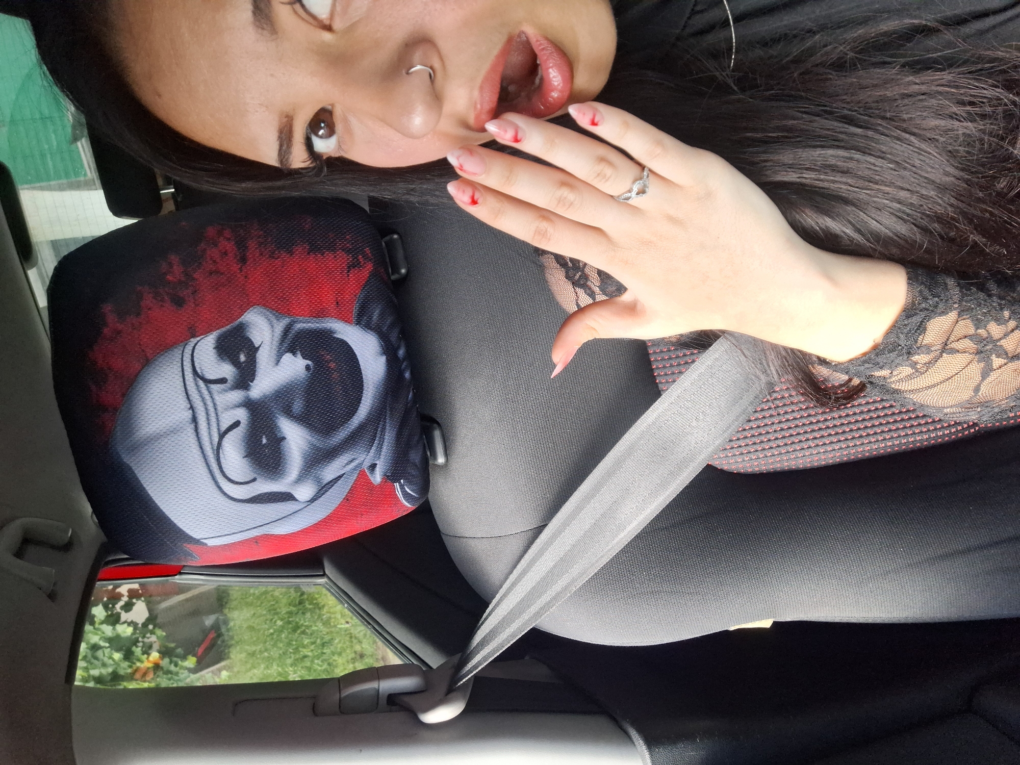 A woman with dark hair, a nose ring, and painted nails with red tips is posing inside a car. She is wearing a black top with lace sleeves. She has a ring on her finger. The seat has a headrest cover with an image of a skull-like figure on a red background. The woman is wearing a seatbelt. The setting is a car interior with a view of greenery and a building visible through the car window. Without additional contextual clues, it is impossible to determine the specific location within Sofia, Bulgaria.