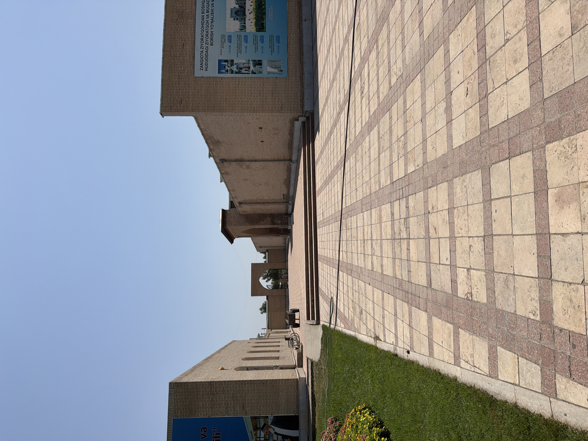 The image shows an outdoor courtyard in Zangiata, Uzbekistan, under a clear blue sky. The foreground features a paved walkway with a geometric pattern of light-colored tiles bordered by red granite. To the left, a strip of well-maintained green grass is bordered by a colorful flower bed.

Dominating the mid-ground and background are low-lying brick buildings with arched entrances and windows. One building on the left has a large blue banner with text. Another building on the right displays a large informational poster with images and text in Uzbek. In the center, an arched gateway leads to another area. A small cart with two wheels is positioned in front of the stairs leading up to the gateway. A bicycle is leaning against the wall of the building on the left. A person wearing a mask and traditional attire is partially visible on the blue banner. The overall scene conveys a sense of historical and cultural significance, likely a religious or memorial site.