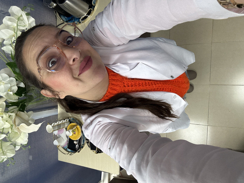 A high-angle photograph depicts a female subject in an indoor environment, identified as Meknes, Morocco. The individual is attired in a white lab coat over a vibrant orange knit sweater and wears rose gold-framed spectacles. Dark hair is pulled back from the face. The subject displays a wide-eyed expression with a slight upward curve of the lips.
The background features a light-colored desk surface against a blue, textured wall. A clear glass vase containing white artificial flowers rests on the desk. To the left, a decorative metal tray holds several small, ornate cups; some white cups are stacked, and a single yellow cup is visible. A dark-colored bag or case is partially visible below this tray. To the right, another decorative metal tray displays a silver electric kettle. The floor is composed of light-toned square tiles, with beige footwear partially visible at the bottom of the frame.