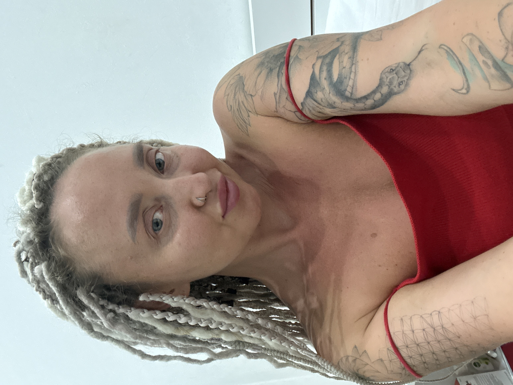A woman with blonde dreadlocks and tattoos on her arms is looking at the camera in a dimly lit room. She is wearing a red top. The room appears to be a bedroom, with a bed and a white wall in the background. There are also some objects on a shelf to the left of the image. The woman is positioned in the center of the frame, and her face is clearly visible.