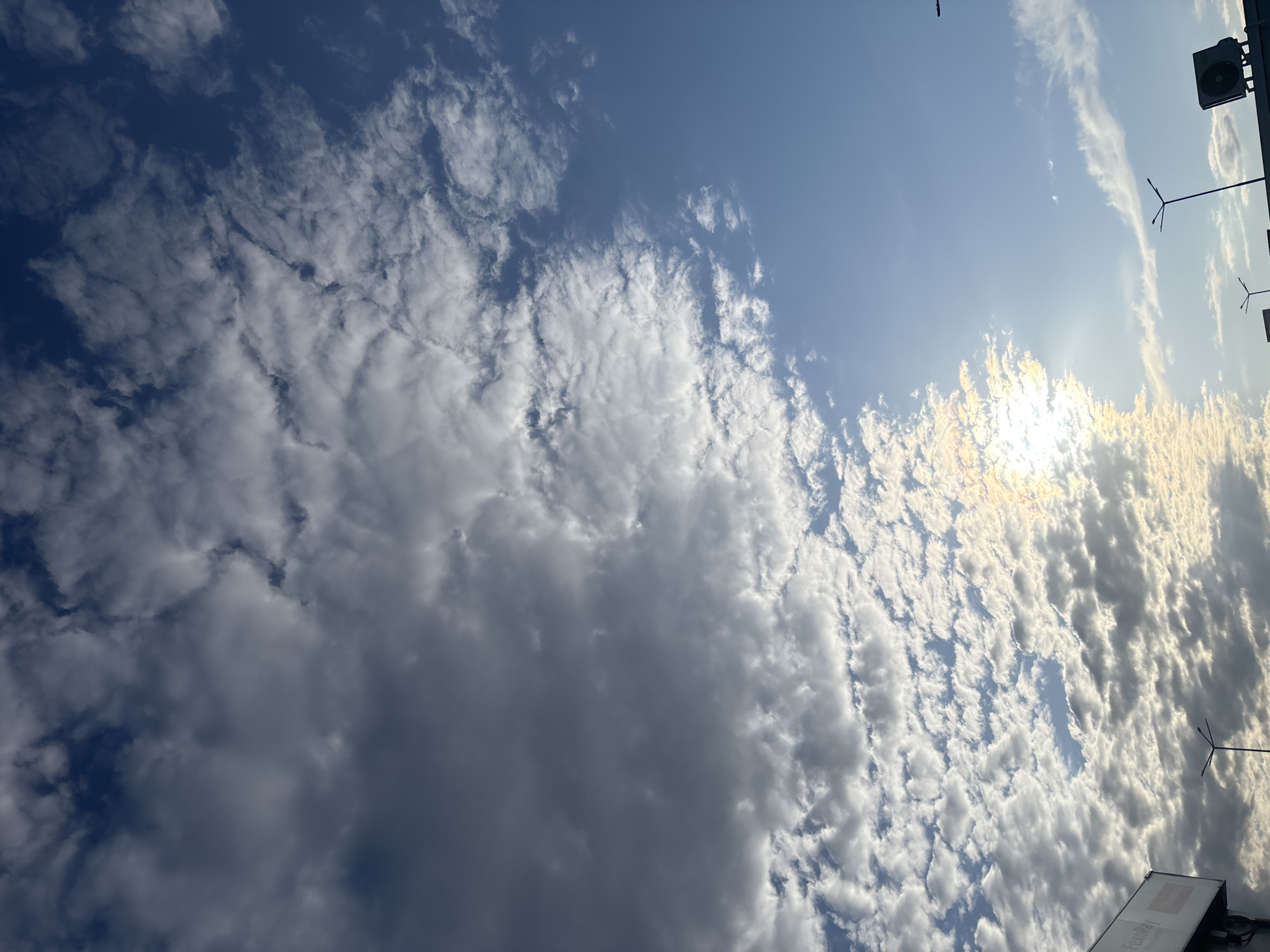 The image shows a sky with many clouds on a sunny day. The clouds vary in density, with some appearing thick and dark while others are thin and wispy. The sun is shining brightly, causing a glare. In the top right corner, there are street lights and a building edge. The location is likely Sofia, Bulgaria, based on visual references such as architectural styles, infrastructure, and cloud types typical of the region. However, the image doesn't provide enough detail to confirm.