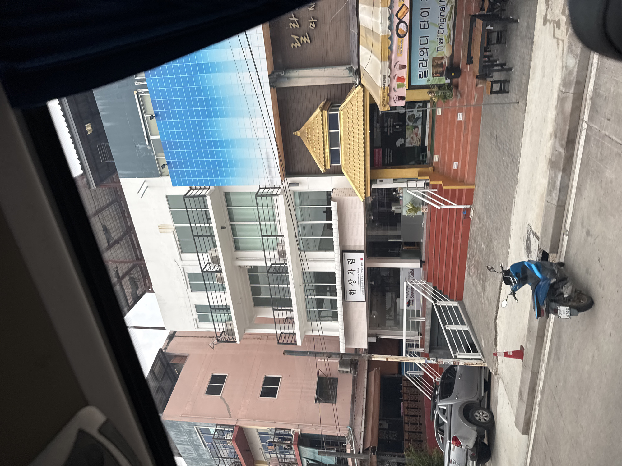 The image depicts a street scene in Bang Phra Subdistrict, Thailand, with a focus on several multi-story buildings. The foreground shows a blue motorcycle parked on the side of the road, with a red traffic cone nearby. A silver pickup truck is parked on the left, facing away from the camera.
The buildings are varied in style and color. On the left is a tall, pale pink building with multiple balconies and windows. Adjacent to it is a white building with several floors, also featuring balconies and numerous windows. One section of this white building is covered by a large, blue, grid-patterned advertisement. To the right of the white building is a brown building with a distinctive golden, multi-tiered roof structure above its entrance, which appears to be a restaurant or shop. This building also has an awning with yellow and white stripes. Signs in Korean and Thai are visible on the buildings, suggesting a mix of businesses.
The street itself is paved, with a sidewalk separating the road from the buildings. The environment appears to be a commercial or mixed-use urban area, with evidence of shops and possibly residential units. The sky is not clearly visible, but the overall lighting suggests it is daytime.