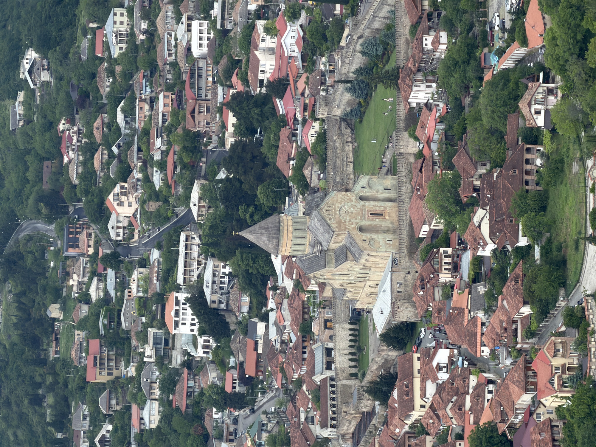 The image shows an aerial view of a dense urban landscape with a mix of residential buildings and green spaces. The buildings have varied architectural styles and rooftops, with red clay tiles being a prominent feature. The density of the houses suggests a closely-knit community. A prominent, large stone building with a cone-shaped roof is in the center of the image. A green lawn stretches parallel to one side of the stone building and people can be seen walking on the lawn. The surrounding environment contains trees that create a natural buffer to the built areas. The city is Mukhatgverdi, Georgia.