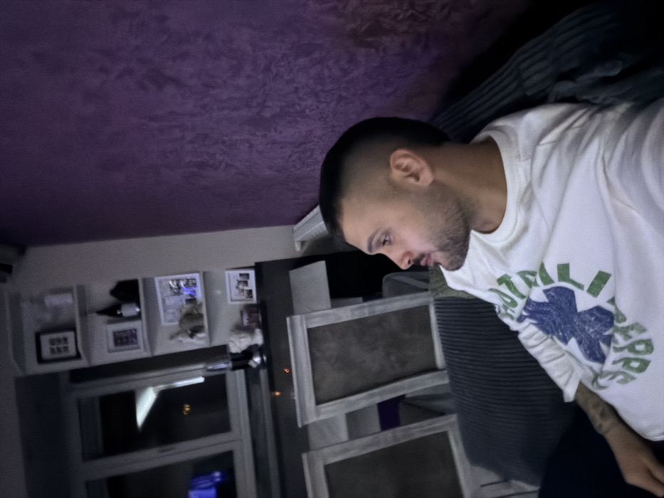 An indoor scene in Sofia, Bulgaria, depicts a male subject seated on a dark, ribbed sofa. The man, seen in profile facing right, has a short fade haircut and a trimmed beard. He wears a white long-sleeved shirt with a blue star-like emblem and green text on the chest, and visible tattoos on his left forearm. His mouth is slightly ajar. To the immediate left of the subject, two framed items with light-colored borders lean against a dark surface. In the background, a large window reflects interior lights and reveals a dark exterior, indicating night. Below the window is a dark surface, above which a series of light-colored wall shelves are mounted. These shelves hold various decorative objects including framed pictures, bottles, a white bust, and a lamp. A small orange light source is visible between the leaning frames and the window area. The wall on the right side of the image features a textured, deep purple finish. The room is dimly lit by artificial light.