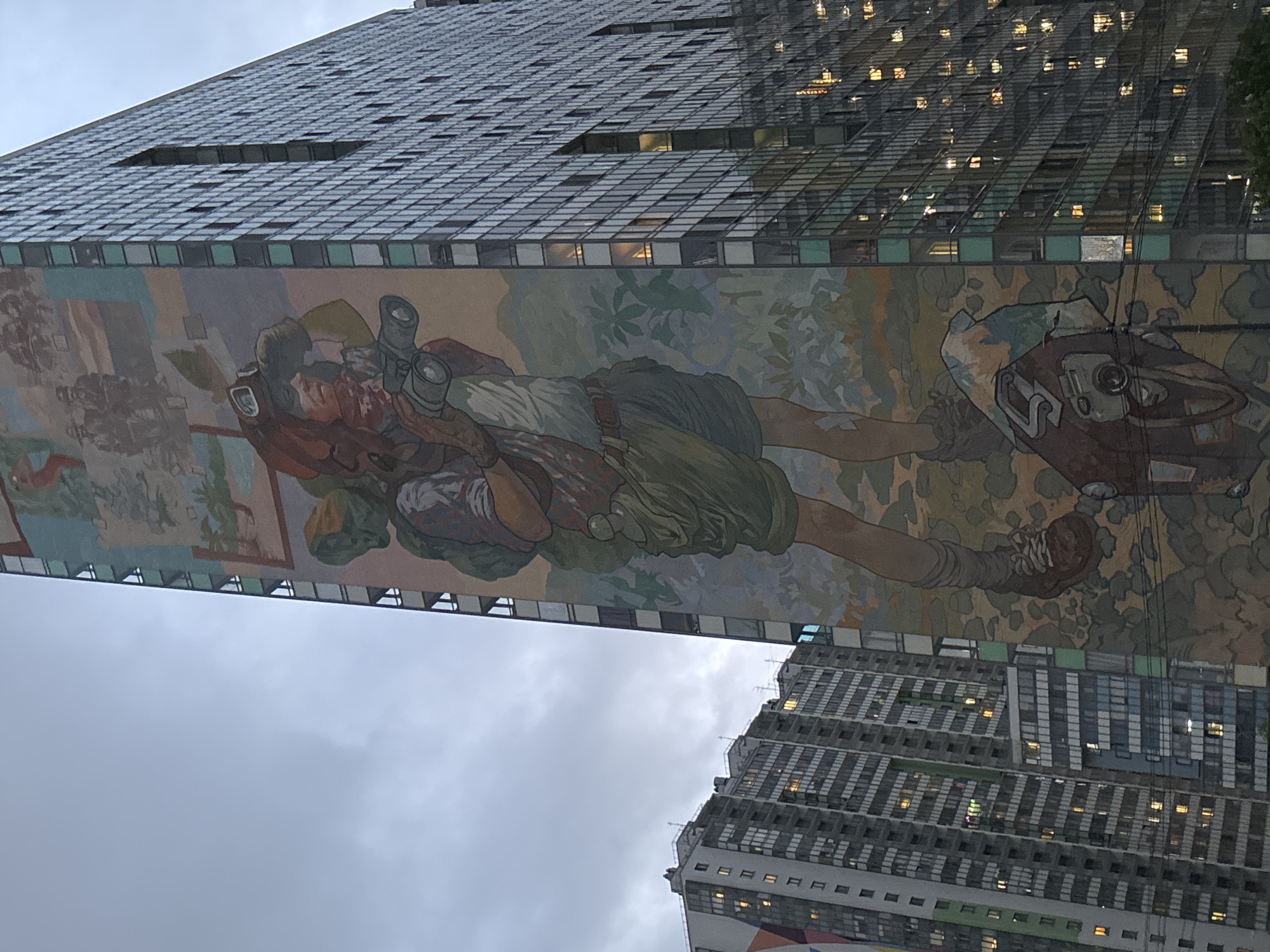 The image shows a mural on a building in St. Petersburg, Russia. The mural depicts a person in what appears to be a historical or fantasy setting, possibly a traveler or explorer. The building the mural is painted on has a modern, grid-like facade with numerous windows. Another tall building with similar architectural details is visible in the background, partially obscured by the mural-bearing building. The sky is overcast, suggesting a cloudy or rainy day.