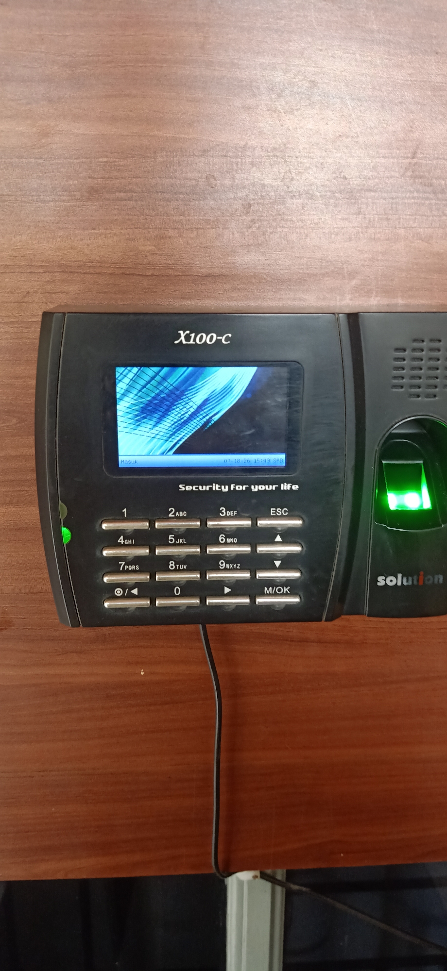 A black fingerprint scanner device, branded "X100-C" and "solution", is mounted on a wooden surface. The device features a small screen displaying abstract blue patterns and text which includes "Masuk", a date "07-18-26", a time "15:49", and "SAB", as well as "Security for your life". Below the screen is a numerical keypad with buttons labeled 1 through 9, 0, M/OK, ESC, and directional arrows. A green illuminated fingerprint sensor is located on the right side of the device. A black wire extends from the bottom of the device and hangs down. The scene is likely within a business or office setting in Palohgadeng, Indonesia, based on the device's functionality and the context provided.