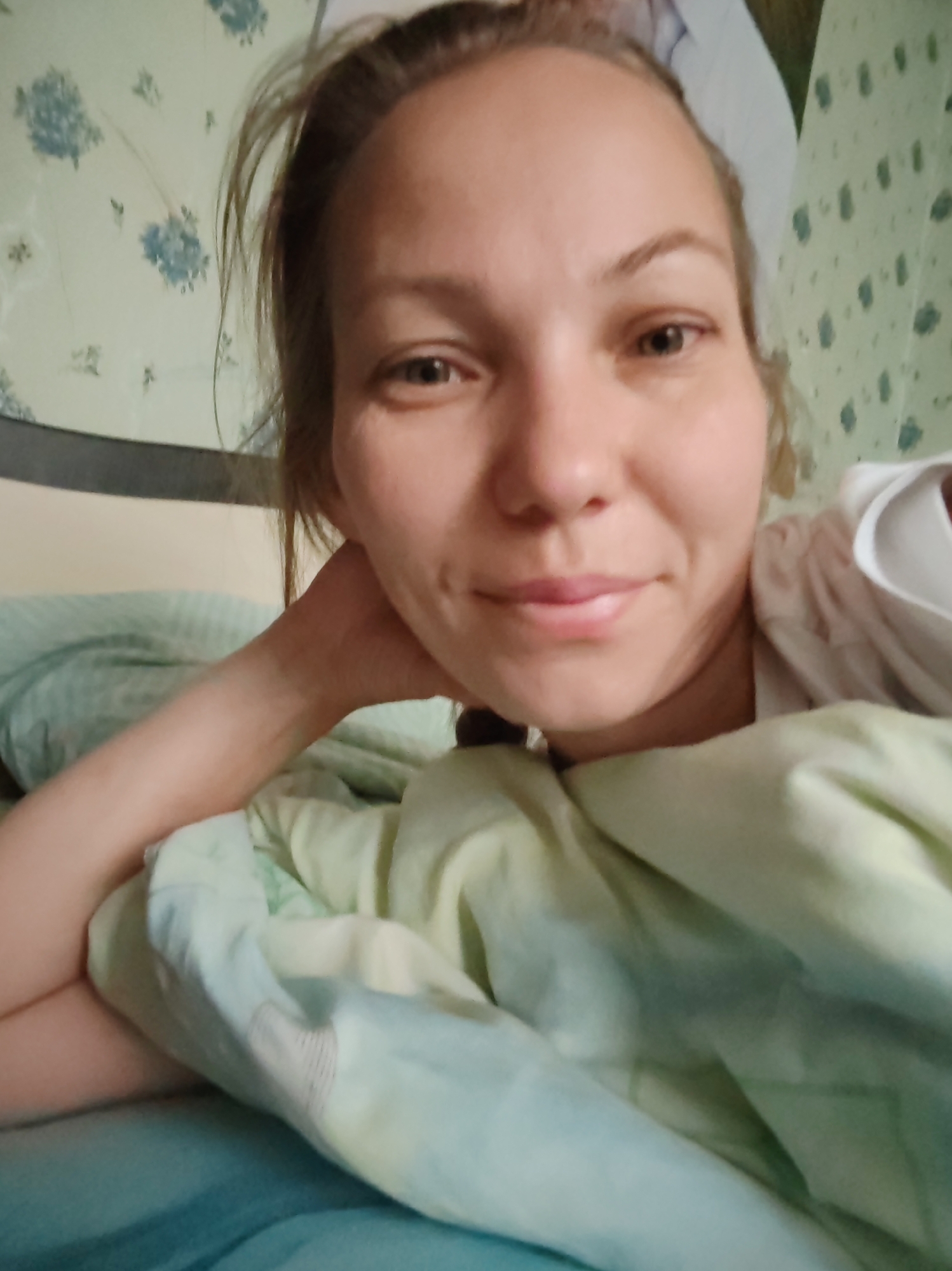 A woman with light brown hair and fair skin is lying in bed. She is looking at the camera with a slight smile and has her left hand supporting her head. She is wearing a white t-shirt. The bed is covered with light green and blue bedding. The background appears to be a wall with a light green floral pattern. The image quality is poor, with low resolution and motion blur. The location is not identifiable from the image.