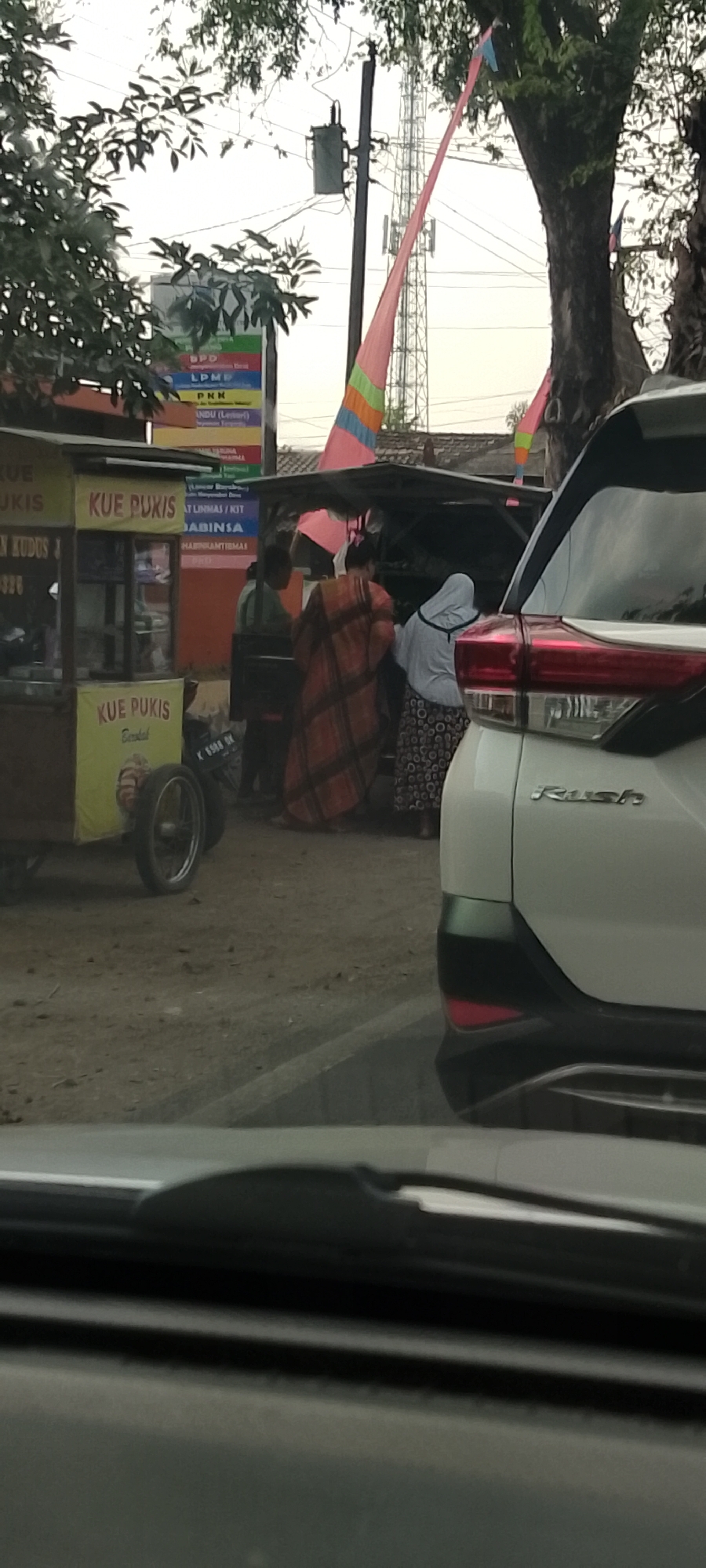 The image captures a street scene in Panjang, Indonesia, with a food stall and several people. In the foreground, the rear of a white car is visible. To the left, a yellow food cart with "KUE PUKIS" branding is parked. Several individuals are gathered around a makeshift stall under a canopy. One person is wearing an orange and brown checkered garment, another a light grey headscarf and patterned skirt, and a third person is wearing a light green shirt. In the background, a tall telecommunications tower stands prominently, with colorful banners strung between trees. Electrical poles and wires are also visible. The ground is unpaved.