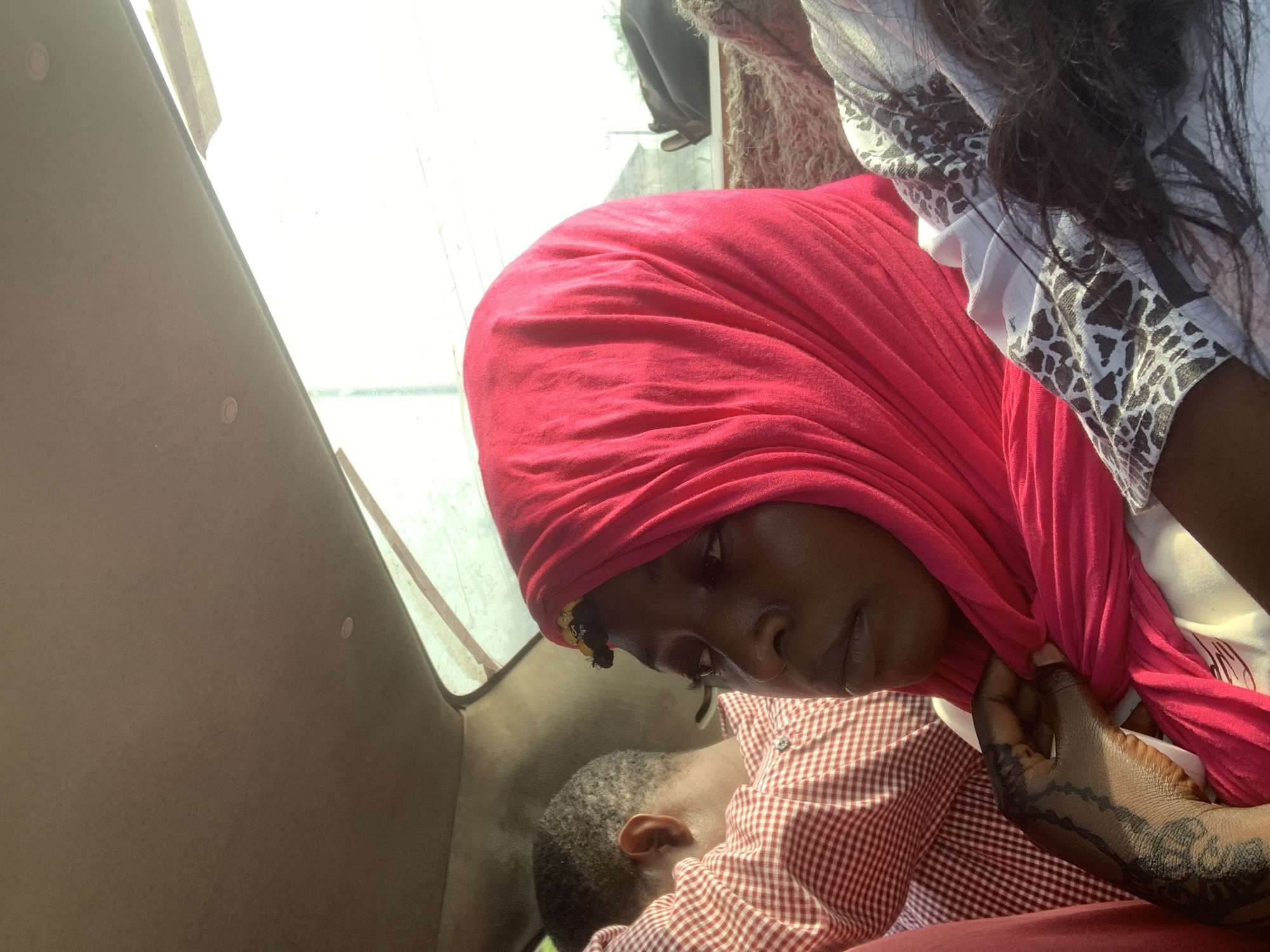 The image depicts a woman wearing a red headscarf. She is looking down with a somber expression. A child with short hair is leaning against her. The woman has intricate henna on her hand and is adjusting her scarf. Another person, wearing a white patterned shirt, is visible in the upper right. The setting appears to be inside a vehicle, indicated by the interior ceiling and window frame. The image was taken in the city of Jiru, Nigeria.
