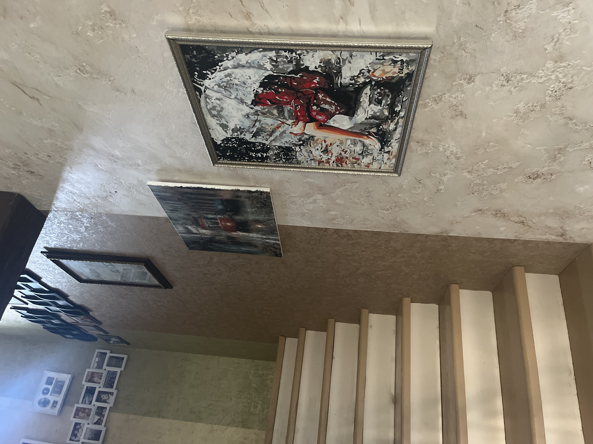 The image shows an interior staircase with several framed pictures and paintings displayed on the wall. The staircase has light-colored treads and risers. The wall to the right of the staircase is textured with a light, mottled finish. A framed painting depicting a woman with an umbrella in the rain is prominently displayed on this wall. To the left of the staircase, a wall with a greenish textured finish features a collection of smaller framed photographs. Further up the wall, a larger framed black and white image is visible. The lighting suggests an indoor setting, possibly a home or building. There are no people visible in the image, and no specific details about the city of Vladimirovka, Russia are present within the visual content.