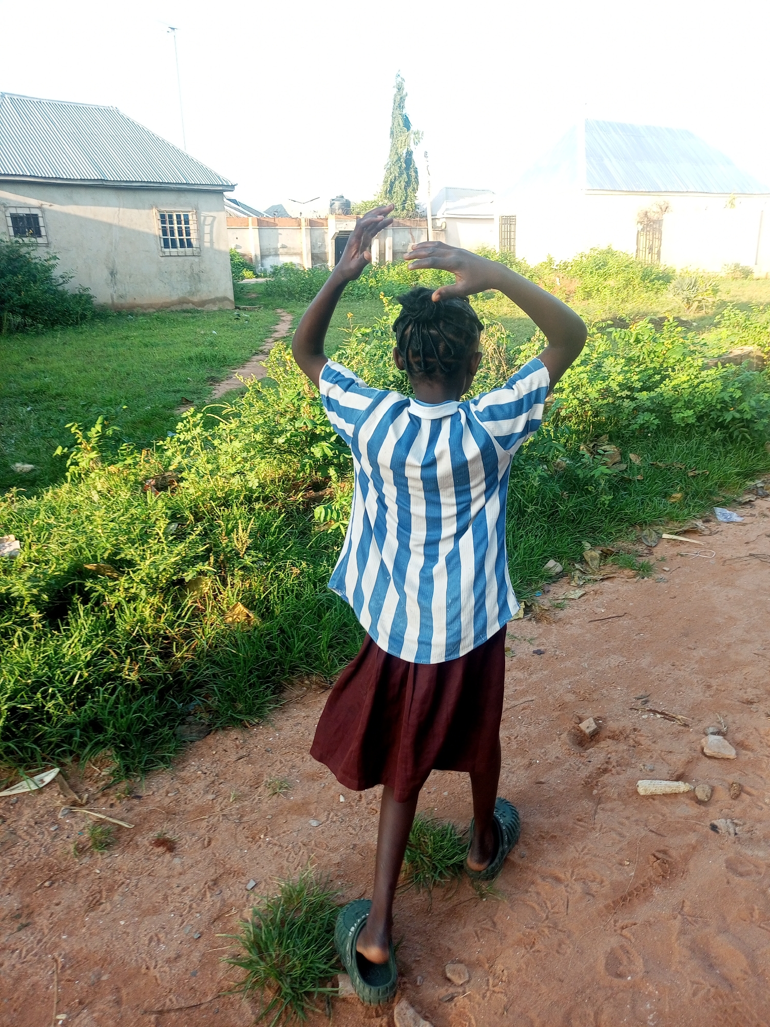 In Tempure, Nigeria, a young girl with dark braided hair, wearing a blue and white striped t-shirt and a dark red skirt, stands with her back to the camera. She has her arms raised above her head, with her hands touching. She is wearing black flip-flops. The girl is standing on a dirt path next to a grassy area. In the background, there are several buildings with corrugated metal roofs and light-colored walls. The sky is bright and overcast. The scene appears to be in a rural or semi-rural area.