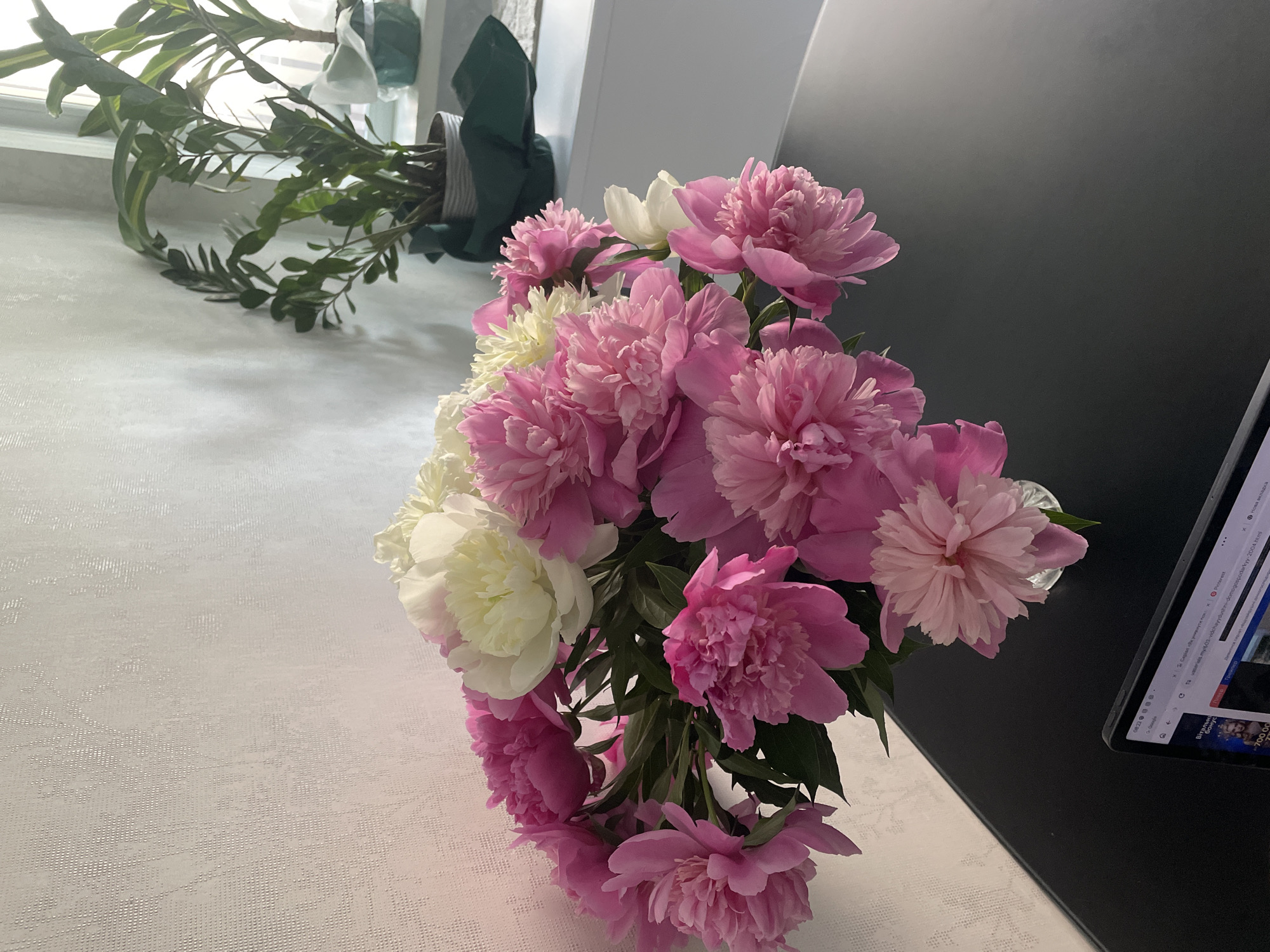 This close-up, high-angle indoor shot captures a vibrant bouquet of peonies set against a bright, domestic backdrop in Rozsoshentsi, Ukraine. The bouquet features numerous full-bloomed peonies, predominantly in shades of pink, interspersed with several creamy white flowers. They are arranged in a clear glass vase, partially visible at the bottom, and placed on a light-colored, textured surface, possibly a tablecloth or countertop, which occupies the left side of the frame.
To the upper left, an indoor plant with slender green leaves is visible, positioned near a window that allows natural light to illuminate the scene. To the right, a dark, reflective surface, likely a table or desk, holds a tablet or laptop. The screen of this device is partially visible, displaying various web elements.
Visible text on the tablet includes the time "08:22", "Google", "Pinterest", and "Нова вкладка" (New tab). Ukrainian text is also present, showing "Серіал «Як уникнути пока ж" (likely part of a TV series title), and a URL "tavaserials.my/603 vidchayydushini-domogospodarkyy-2004.html" which references "Відчайдушні домогосподарки" (Desperate Housewives). Further down, "Вітальни бонус" (Welcome bonus) and "700.00" are visible.
The scene conveys a calm morning atmosphere, blending natural beauty with elements of modern technology in a well-lit living space. No individuals are present, and the primary focus is on the aesthetic arrangement of the flowers.