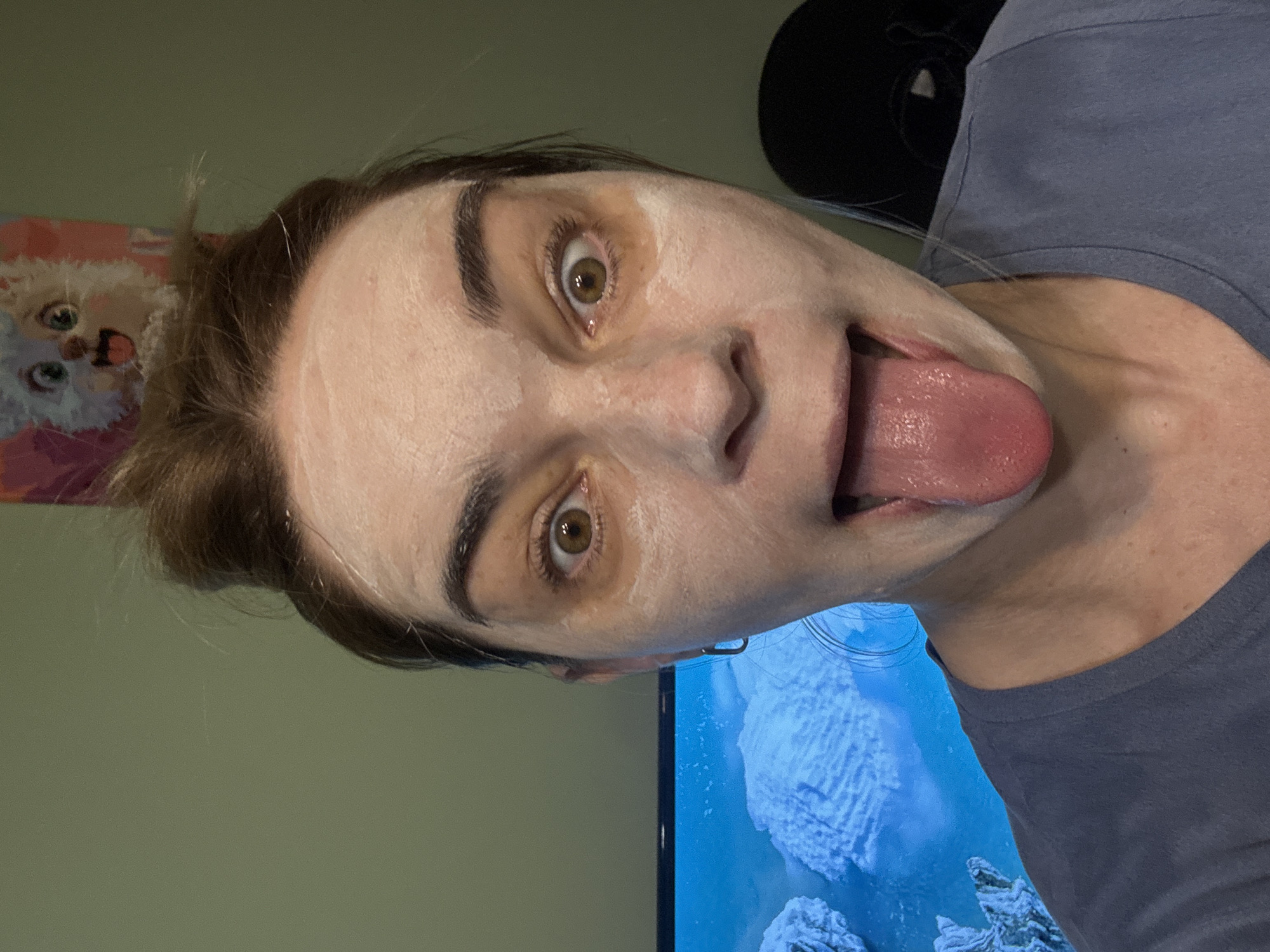 The image shows a woman indoors with a white face mask. Her brown hair is pulled back, and she's wearing a gray t-shirt. She has brown eyes and is sticking her tongue out. Behind her is a green wall, a poster with a cat on it, a black hat, and a television screen displaying a snowy landscape. The location is Unknown, Unknown.